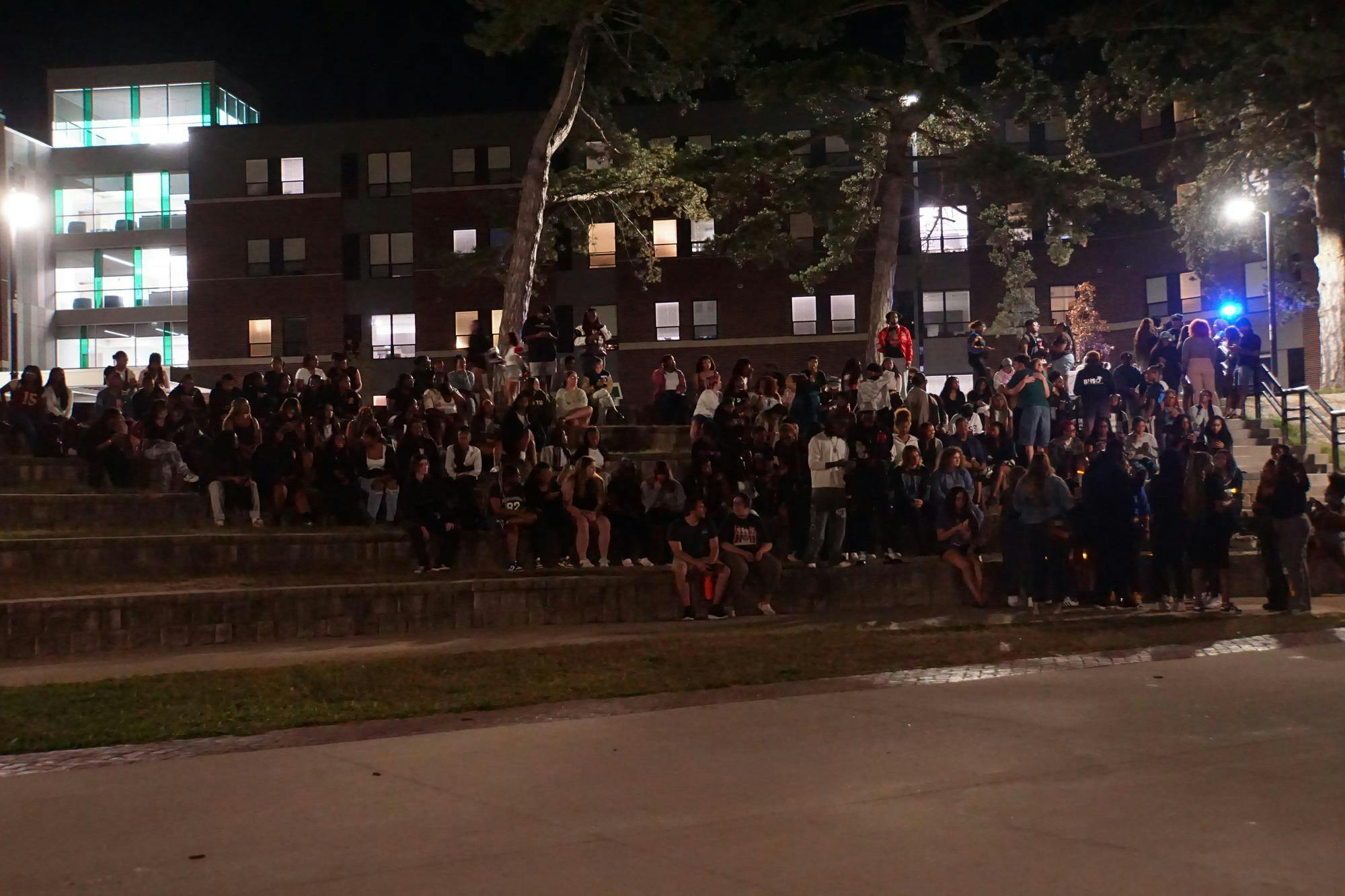 About 100 students sit and stand on the steps of an outdoor amphitheater at night.