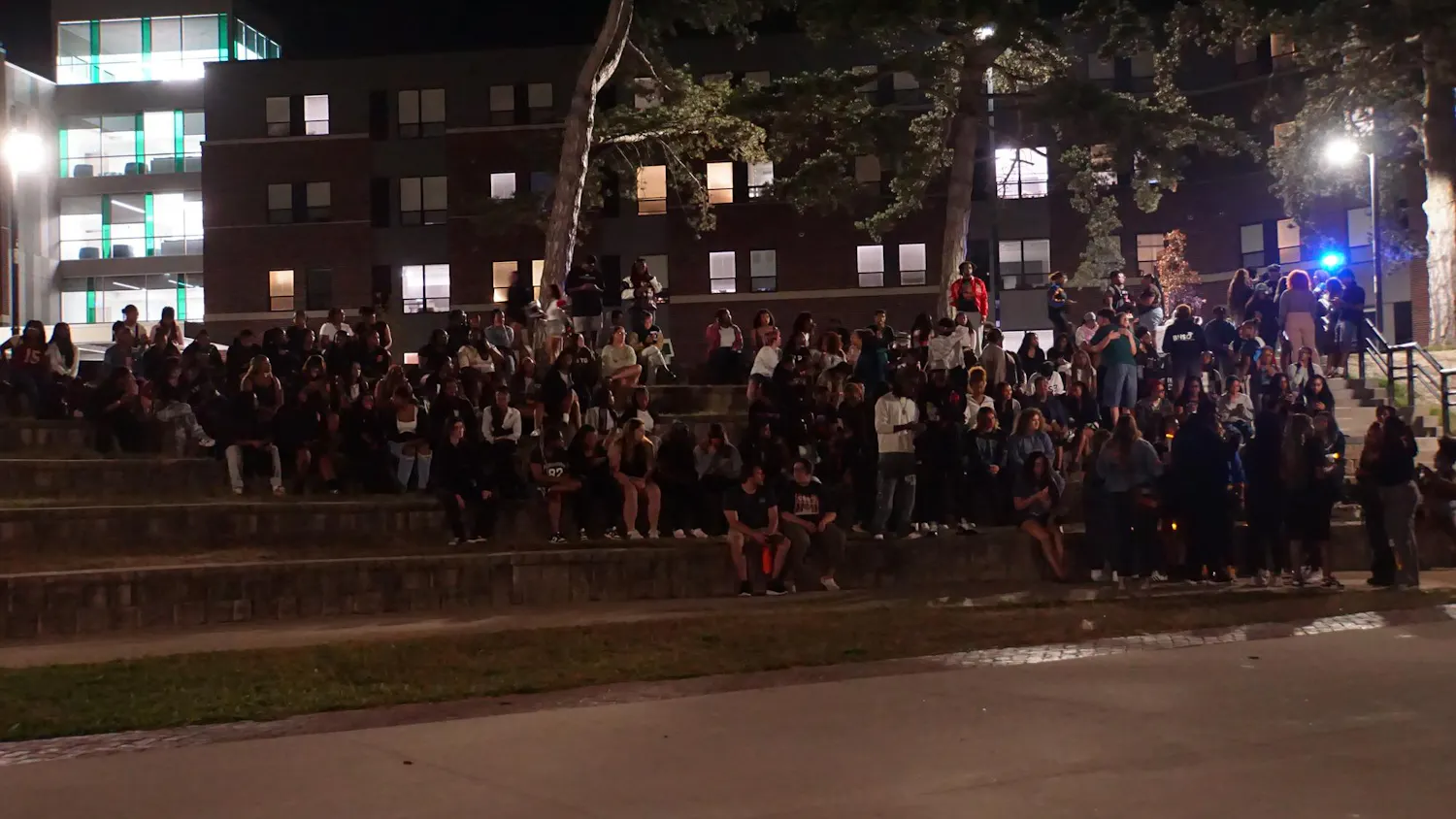 About 100 students sit and stand on the steps of an outdoor amphitheater at night.