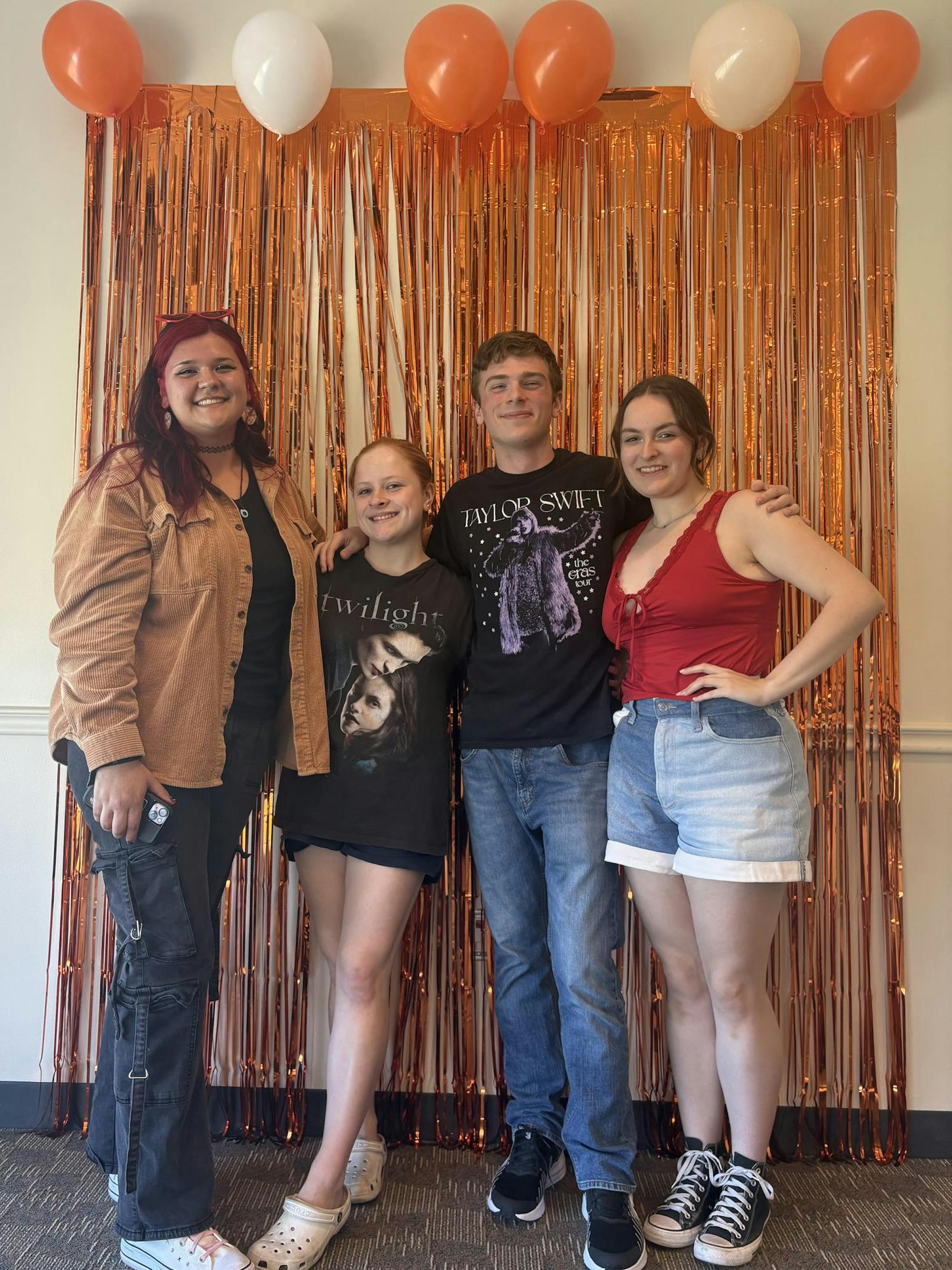 Four students pose in front of a backdrop of metallic orange streamers and orange and white balloons.