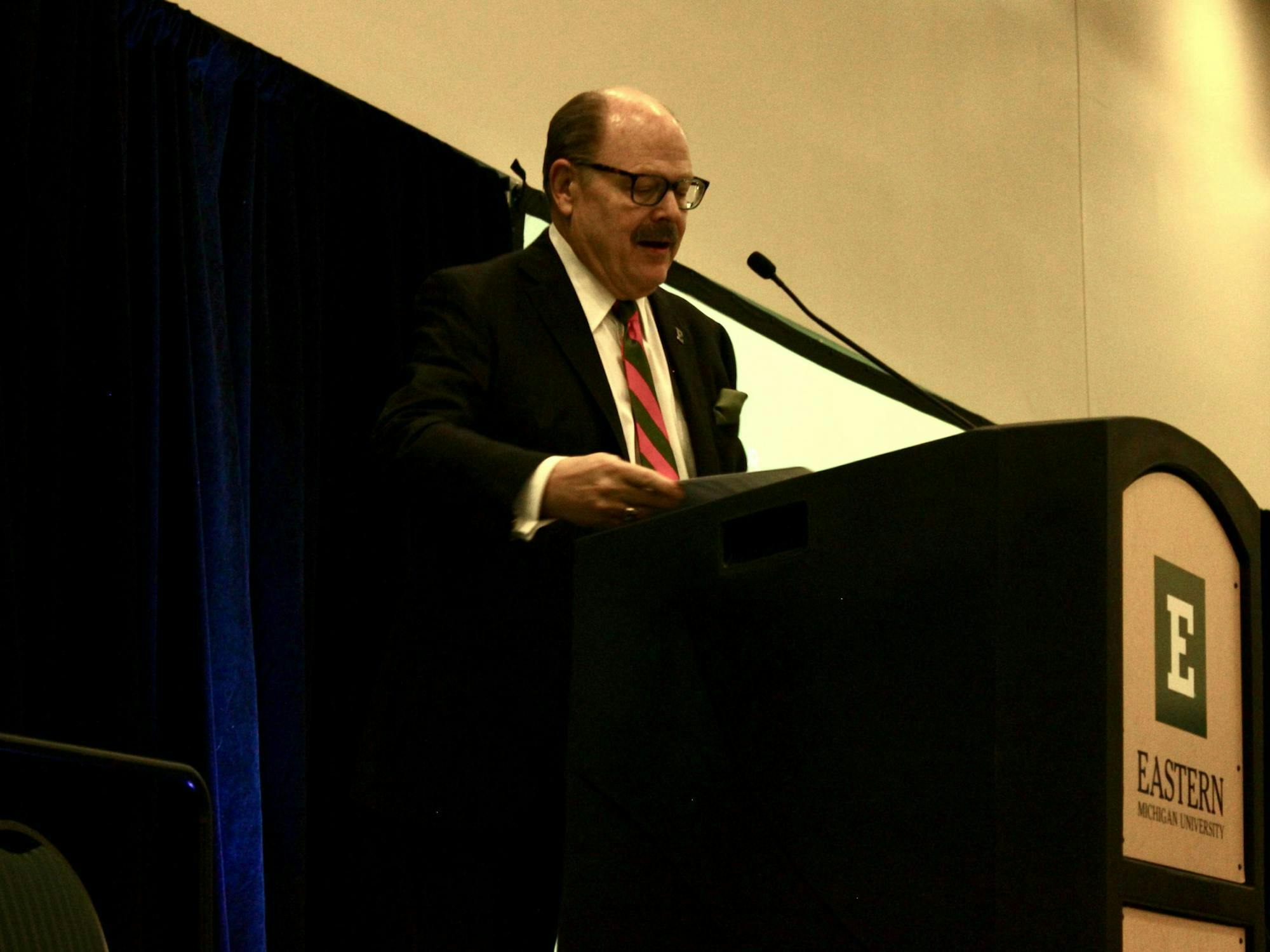 EMU President James Smith stands at a podium with a green and white "E" logo printed on the front.