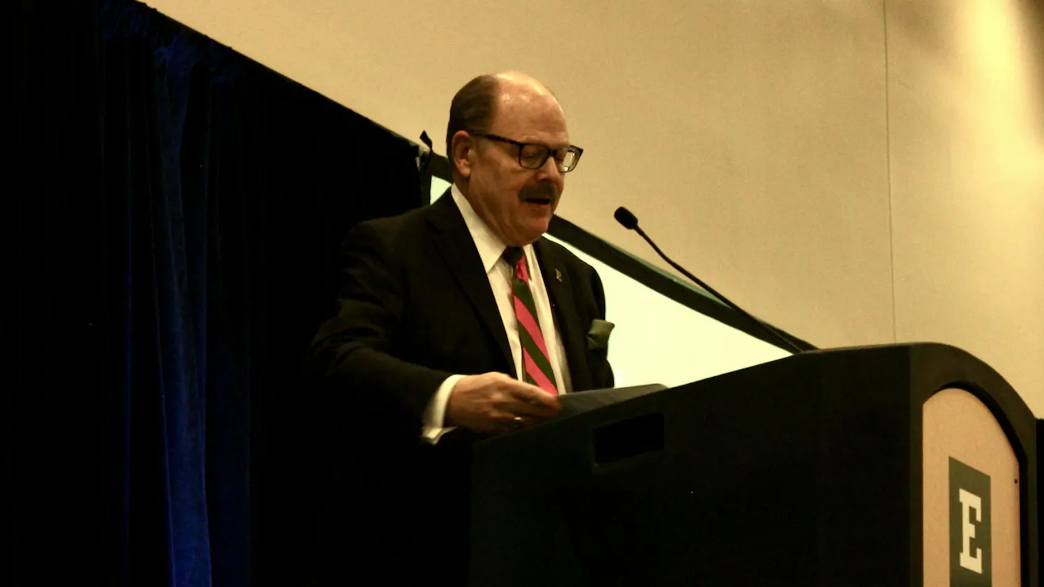 EMU President James Smith stands at a podium with a green and white "E" logo printed on the front.