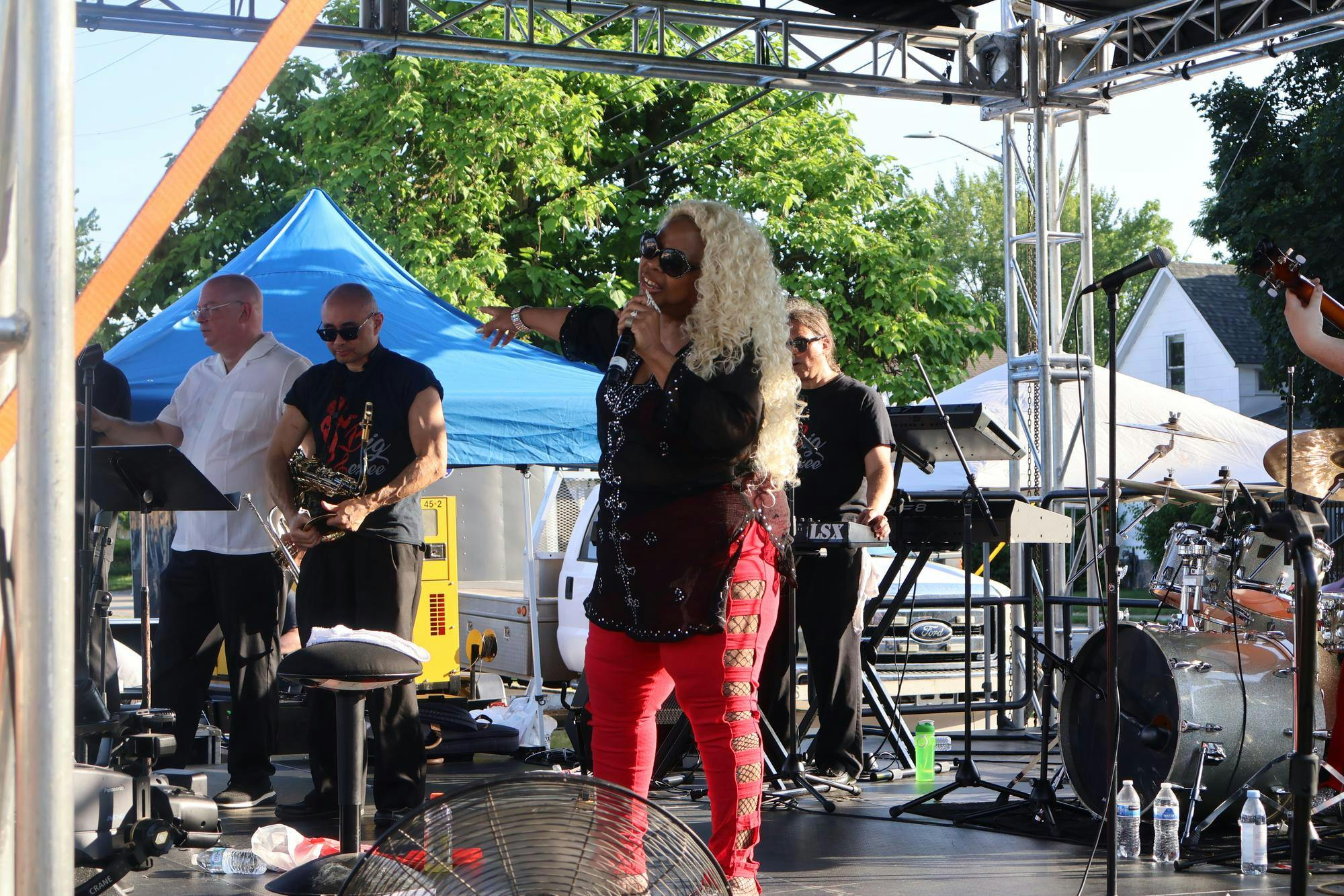 Lady Sunshine has long curly blonde hair, with a bedazzled black blouse and red pants. Band members are seen behind her dressed in black with a saxophone, keyboard, drum set, and guitar. 