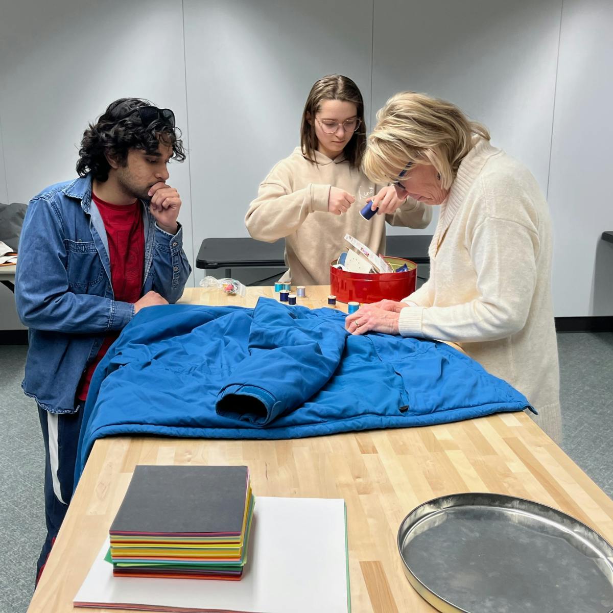 Three people stand at a tan-colored table in the Halle Library Creative Studio on Eastern Michigan University's campus. They are working to repair a blue coat in the Halle Fix-It Hub. 