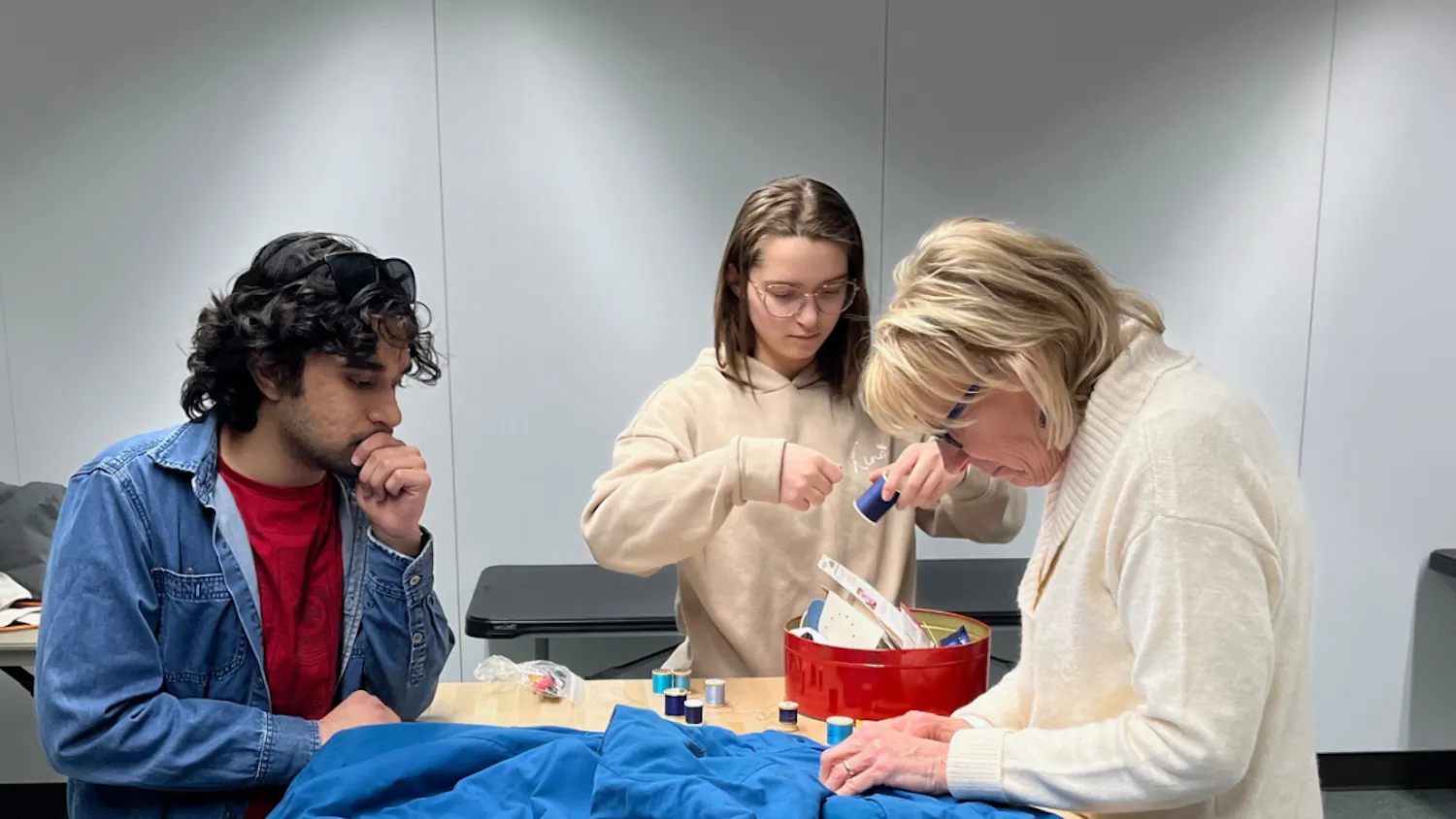 Three people stand at a tan-colored table in the Halle Library Creative Studio on Eastern Michigan University's campus. They are working to repair a blue coat in the Halle Fix-It Hub.