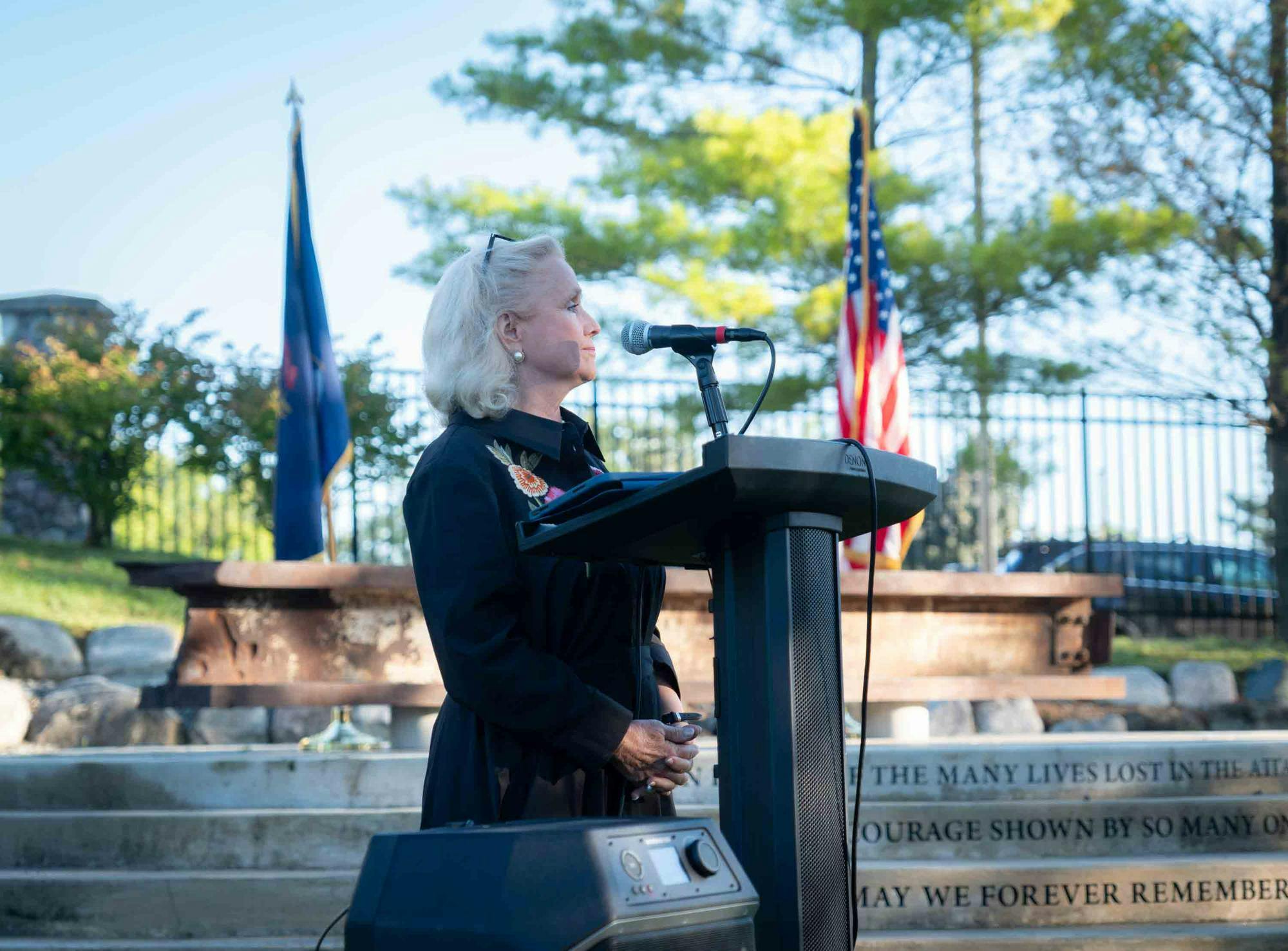 Debbie Dingell stands at a podium outside in front of a memorial on campus comprised of a steel beam from the South Tower of the World Trade Center. A Michigan and United States of America flag are both seen in the background. 