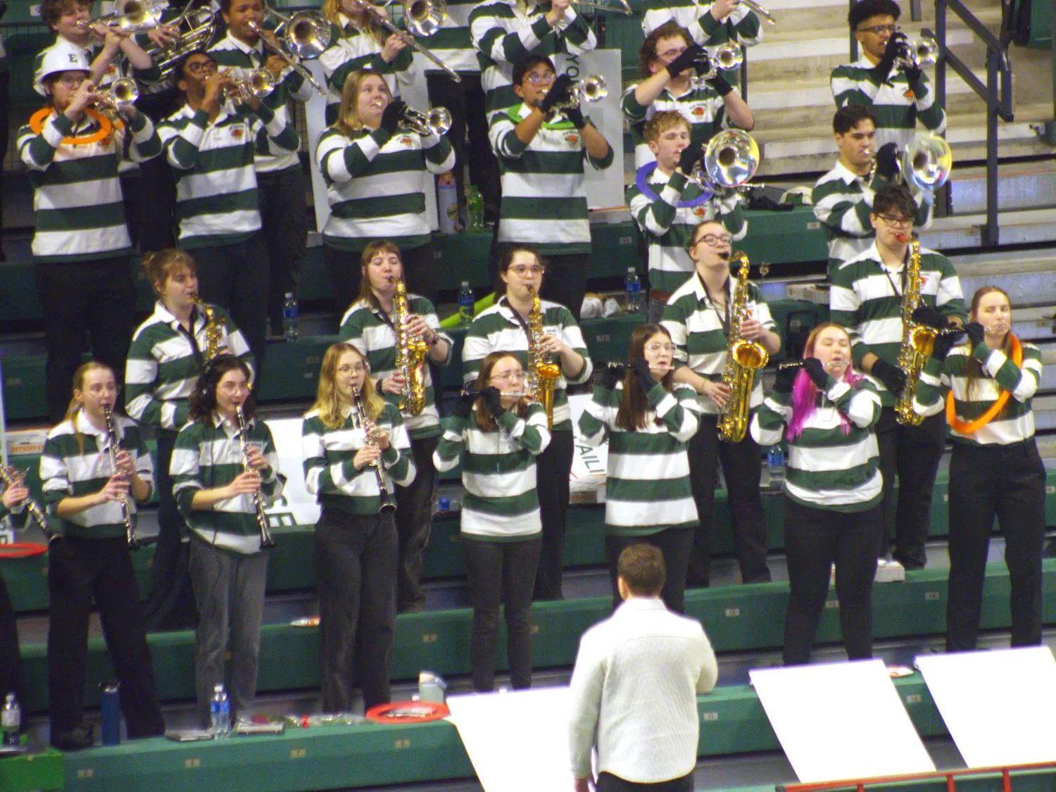 A group wearing white and green stripped shirts stands in the bleachers playing wind instruments.