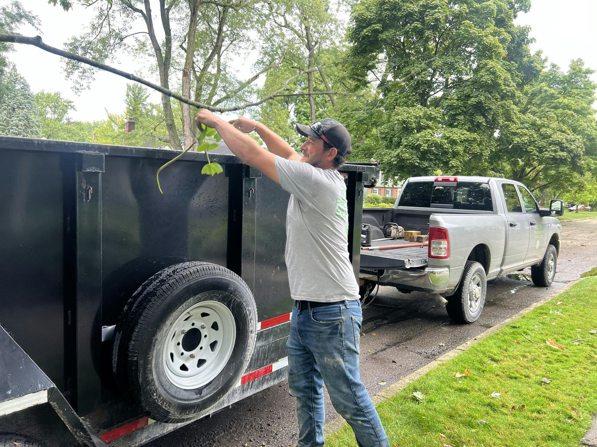 A man throws a tree branch into a trailer behind a pickup truck in a residential neighborhood.