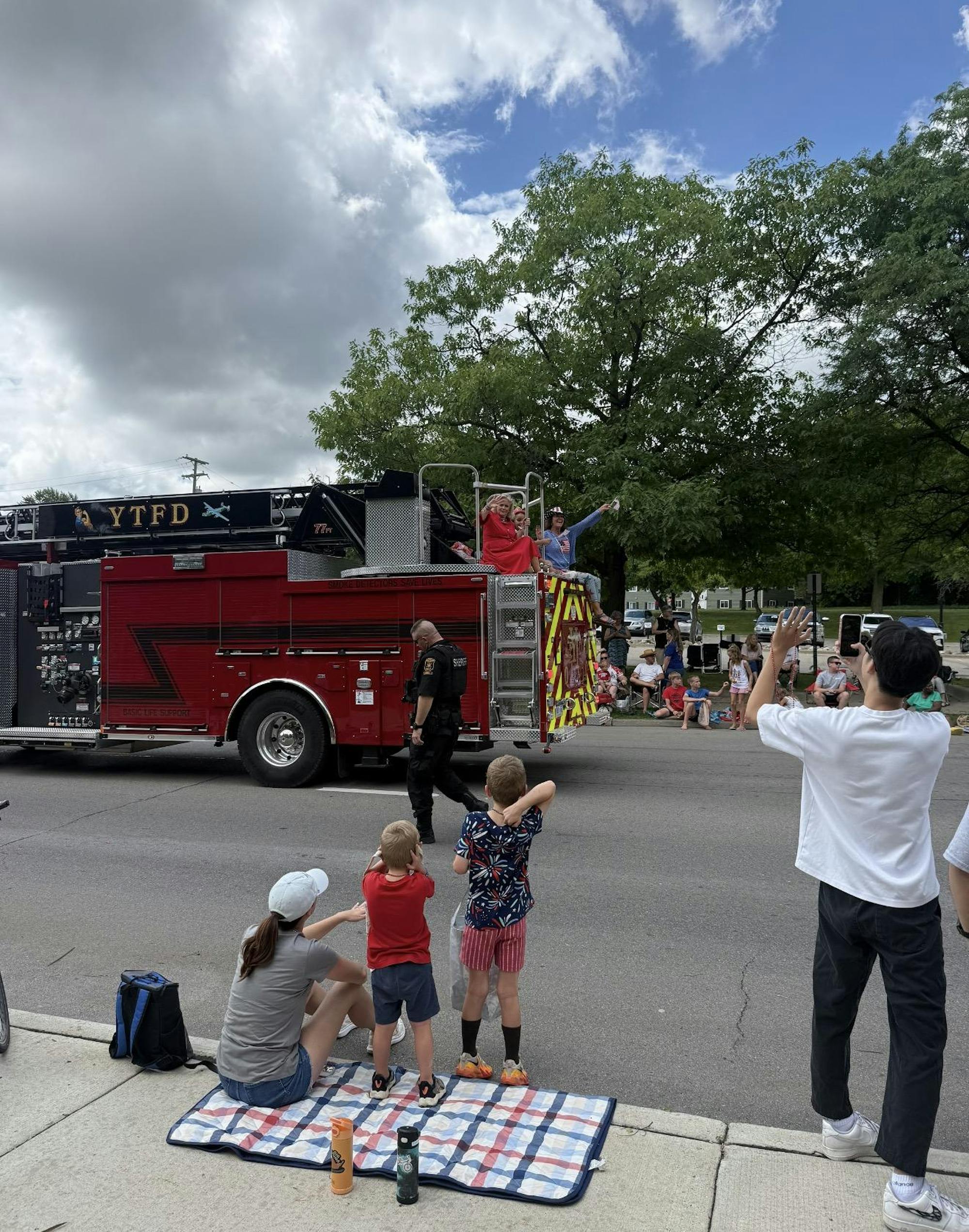 Onlookers stand and sit on the sidewalk and grass while a red firetruck drives by in the parade; on top of the firetruck, Debbie Dingell and other participants wave to the crowd.