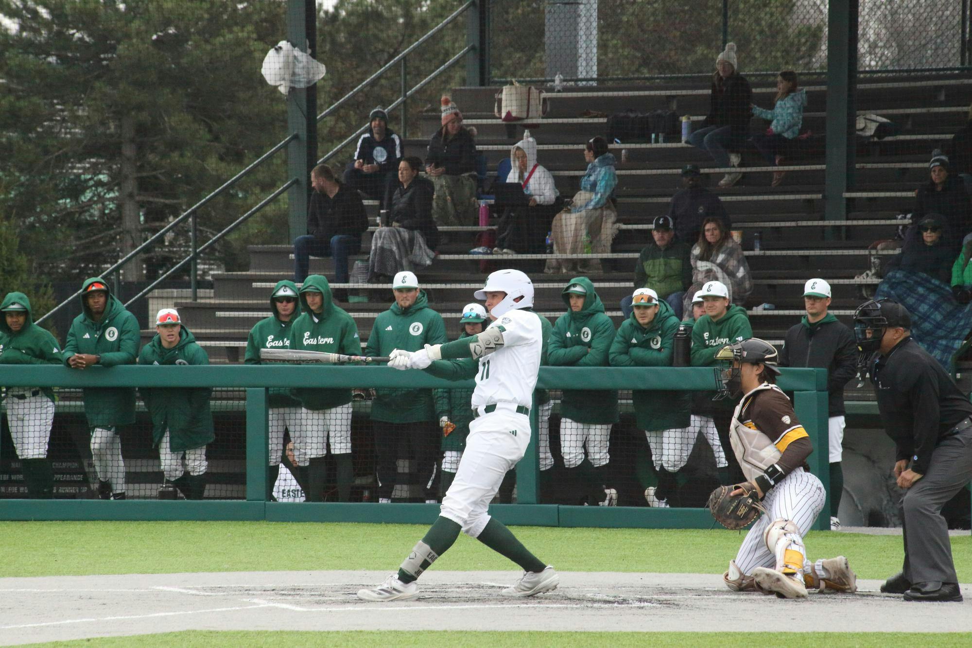 Baseball player mid-swing at outdoor baseball game while teammates watch in the background.