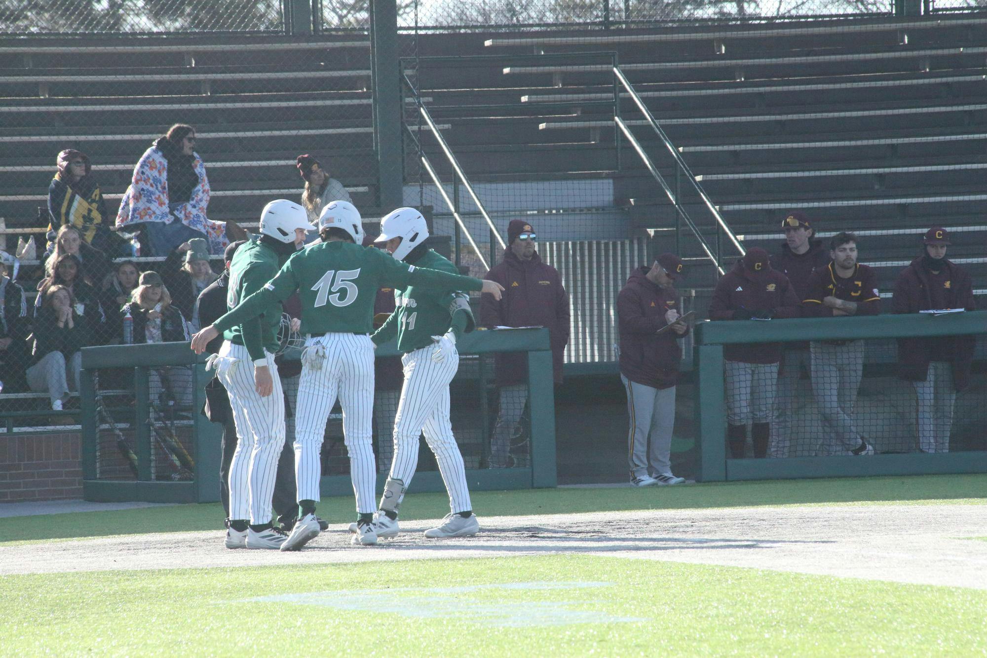 Three baseball players of the same team huddle while spectators watch from the stands in the background.
