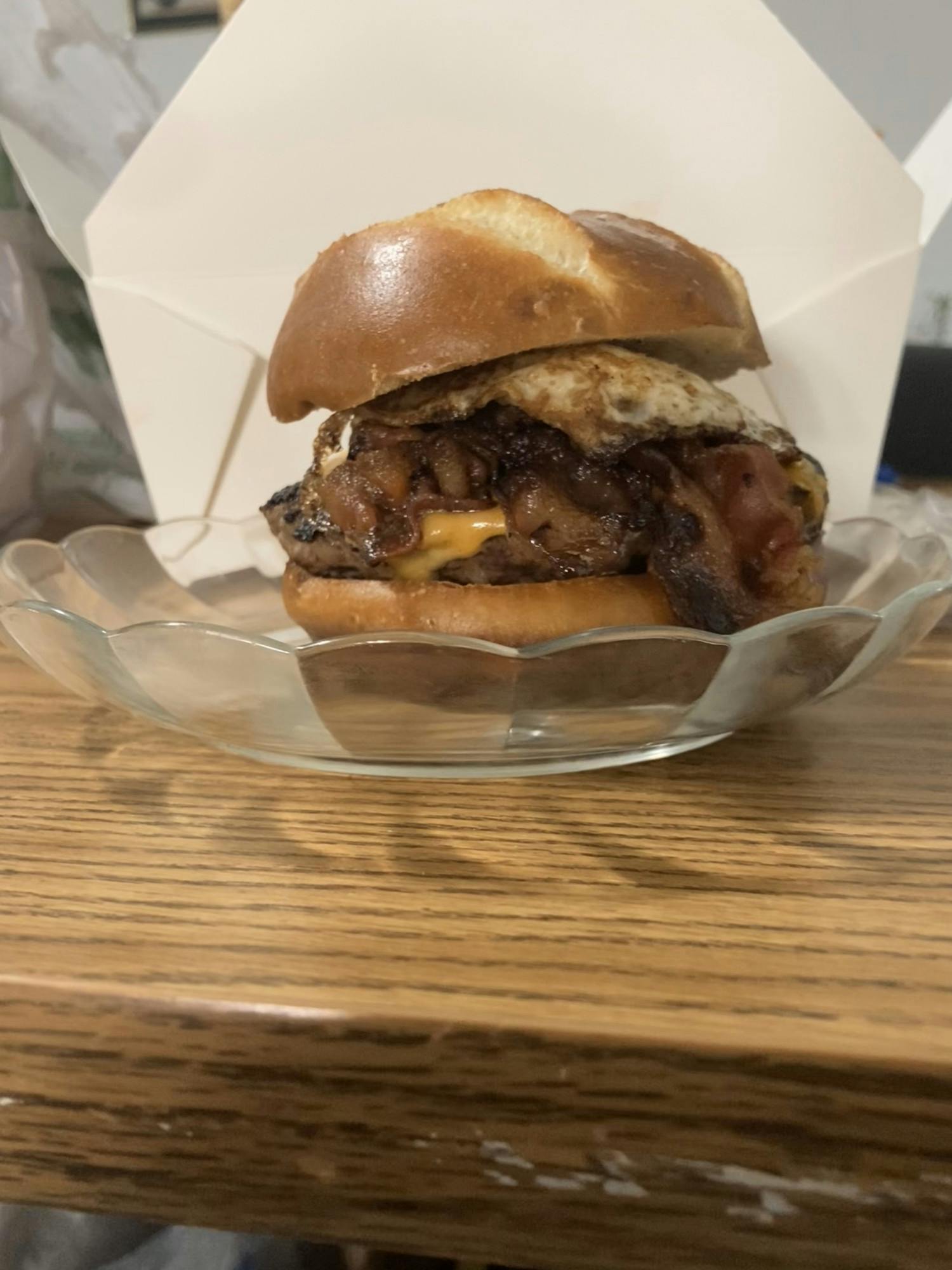 The image shows a juicy-looking burger patty nestled between two burger buns with some bacon and cheese. The burger is on a clear plate with scalloped edges.