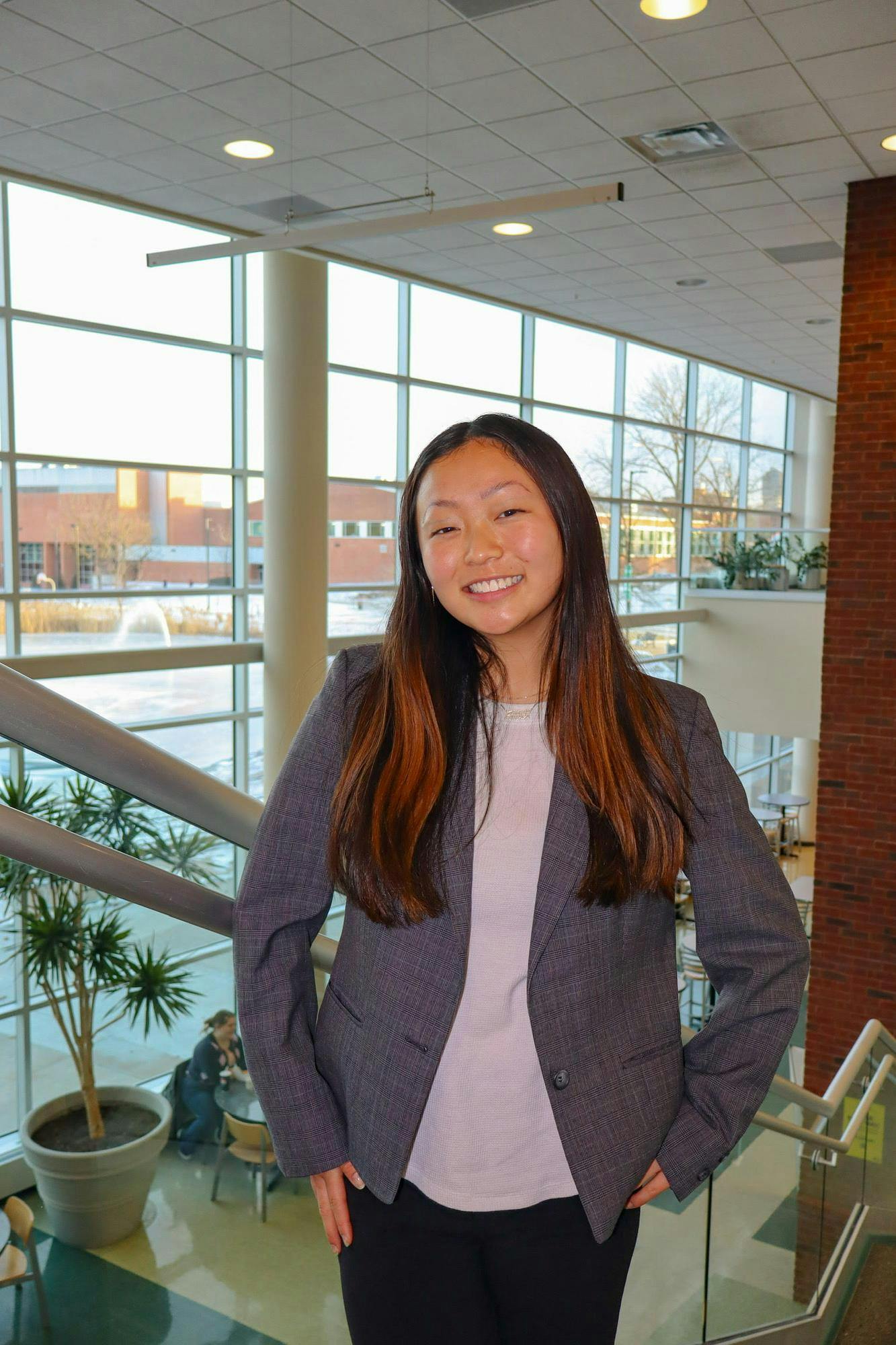 A smiling young woman with long dark hair stands in front of a staircase. She is wearing a dark gray blazer, dark-colored pants, a light gray top and a gold-colored necklace. Behind her is a large window through which a fountain and brick buildings can be seen.