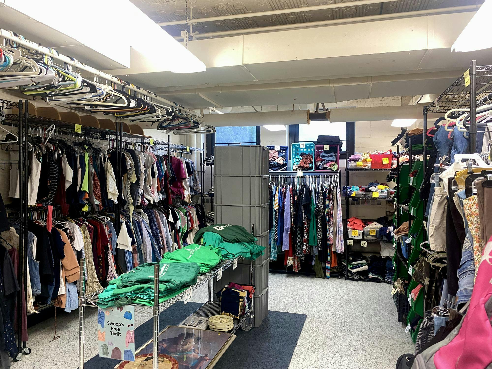 The corner of a room is lined with two-level clothing racks and a table in the center. The racks are filled with hanging clothing.