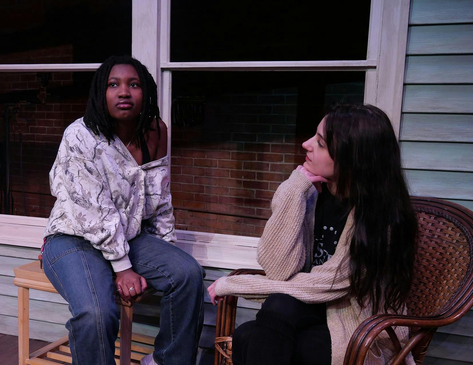 Two actors sit beside one another on a theater set, sat in front of the grey exterior of a house. Claire, who sits on the right, looks at Catherine as she stares off contemplatively. 