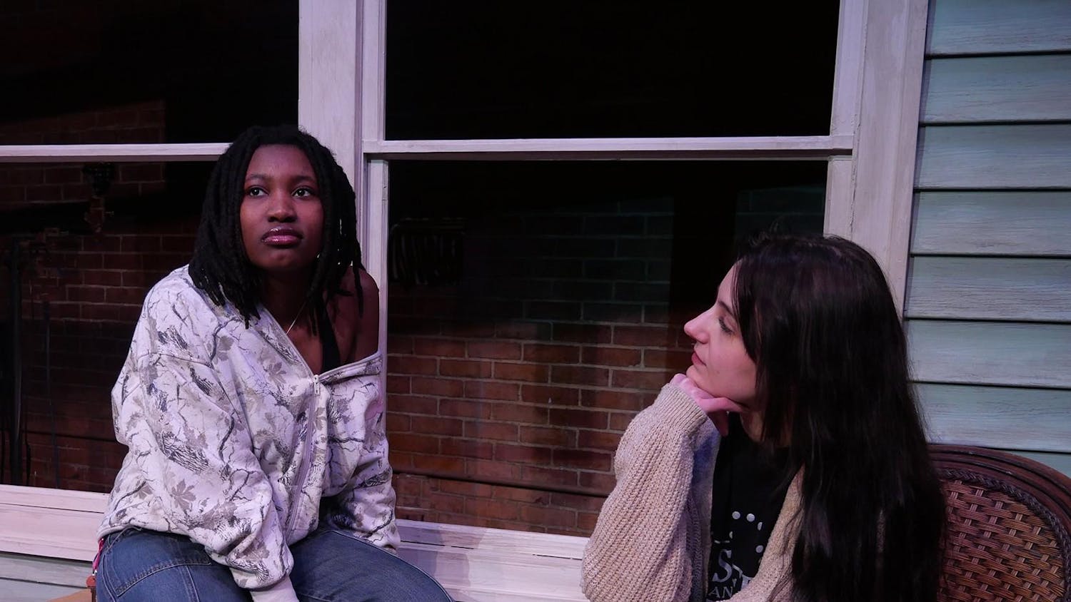 Two actors sit beside one another on a theater set, sat in front of the grey exterior of a house. Claire, who sits on the right, looks at Catherine as she stares off contemplatively.