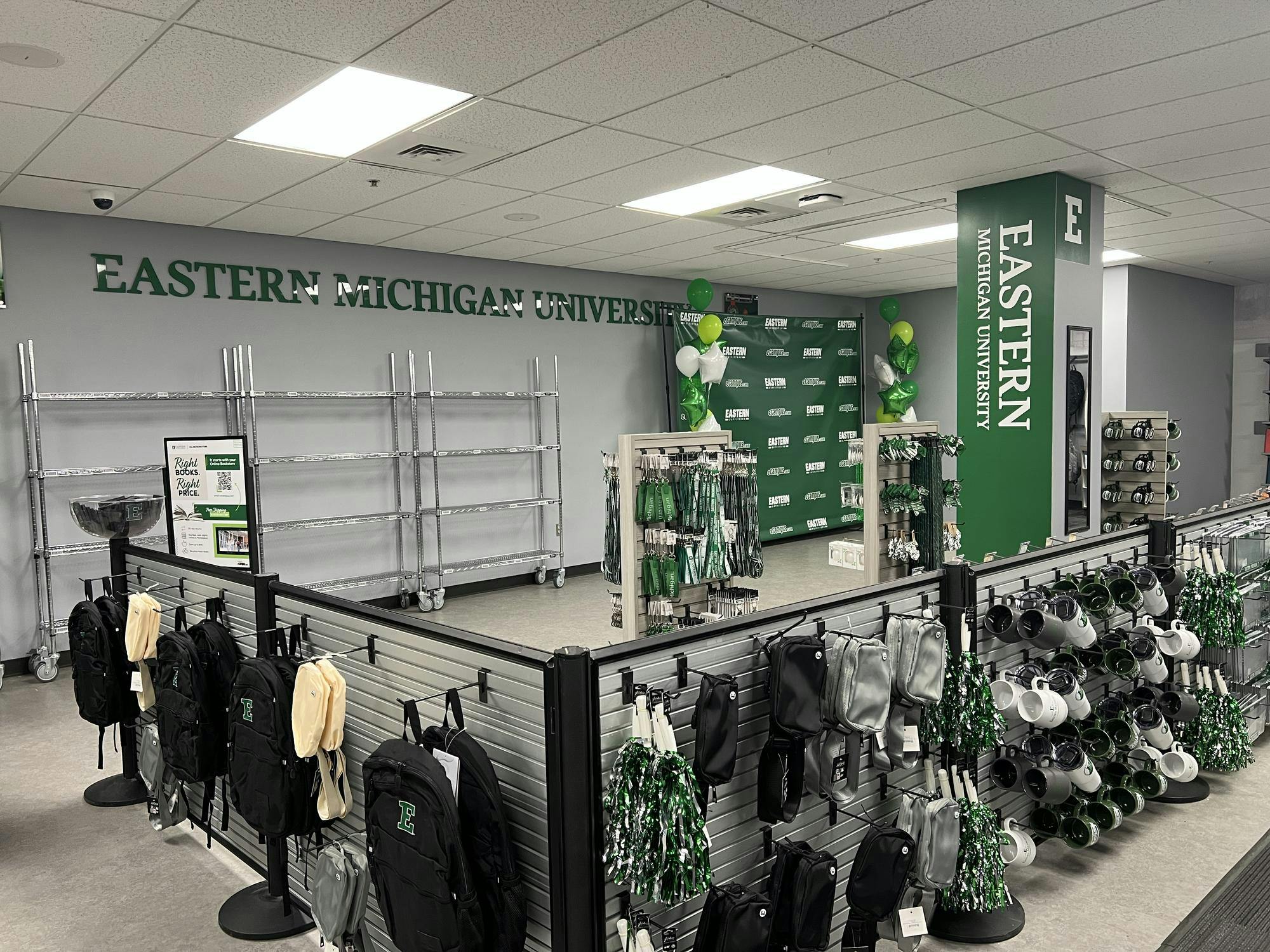 Eastern Michigan University branded backpacks, mugs and other accessories hang on hooks in a store with a gray and green color scheme.