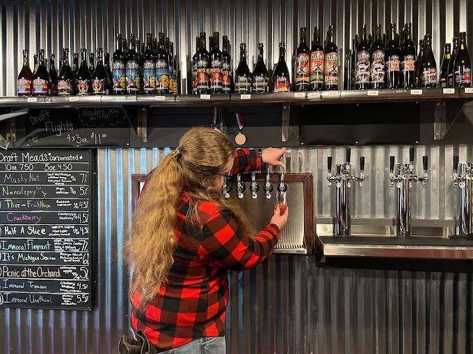A bartender stands before a row of spigots and pours a glass of mead. 
