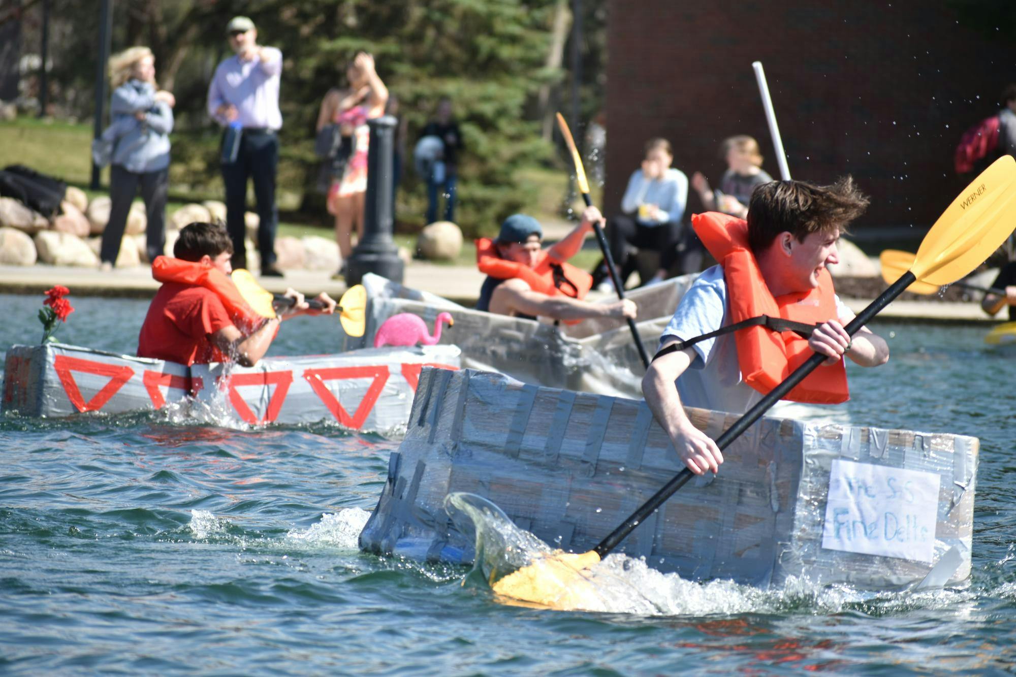 A student rows a handcrafted boat, made with a combination of cardboard, plastic, and duct tape, across a pond. Two other students row handcrafted boats behind them.