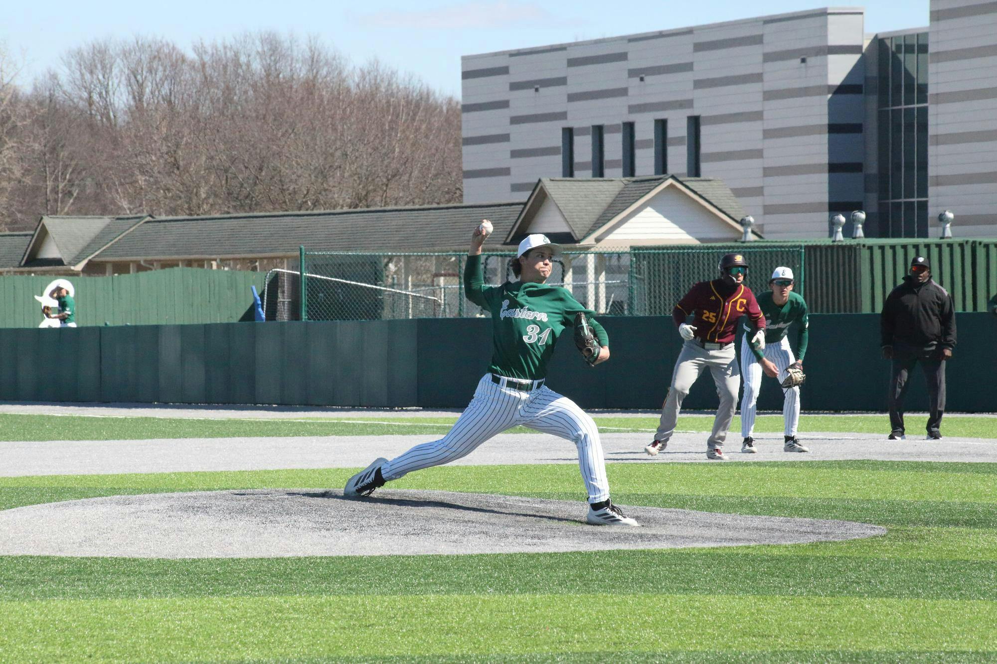 Pitcher mid-toss at outdoor baseball game. Two players stand crouching in the background, one from each team.