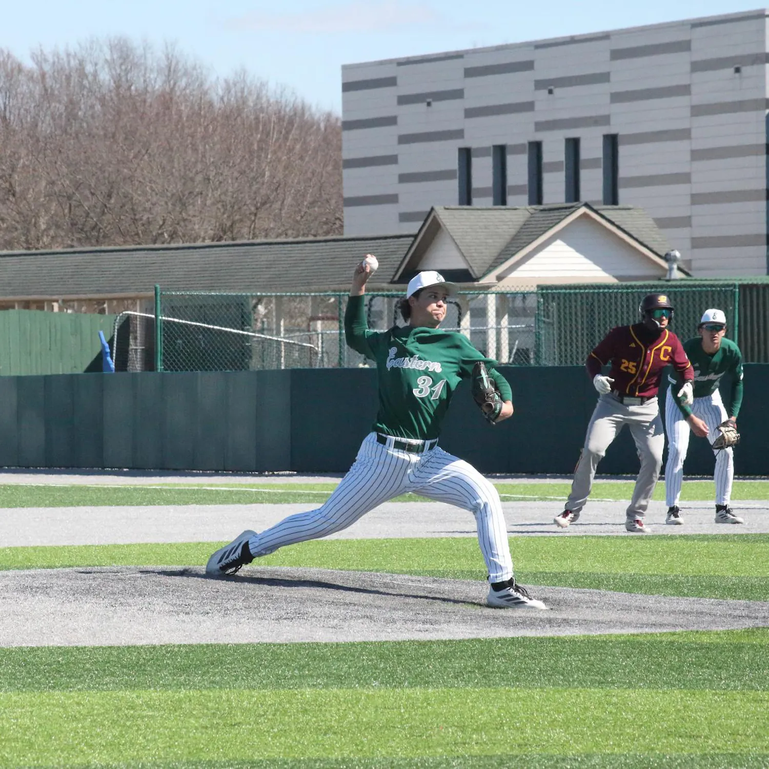 Pitcher mid-toss at outdoor baseball game. Two players stand crouching in the background, one from each team.