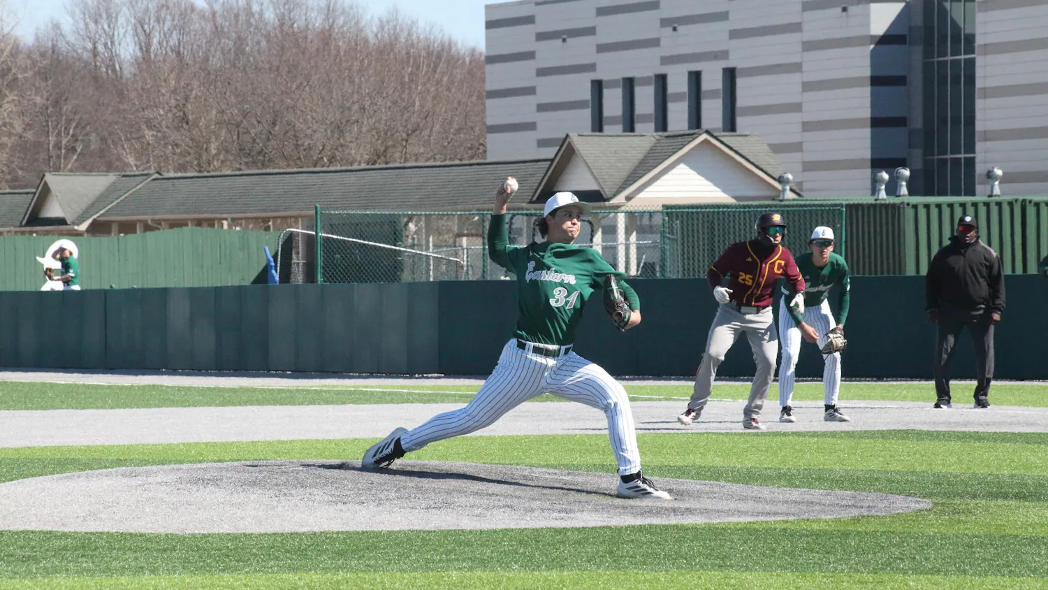 Pitcher mid-toss at outdoor baseball game. Two players stand crouching in the background, one from each team.