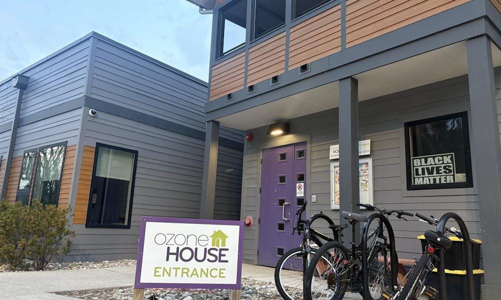 A square-shaped building made of gray-colored wood with a brown-colored panel running across its middle. There are several windows, and the door to the building is purple. In one of the windows is a Black Lives Matter sign. A few bikes are parked in front of the building, and a sign in the foreground says "Ozone House Entrance." Overhead, the sky is a shade of pale blue.