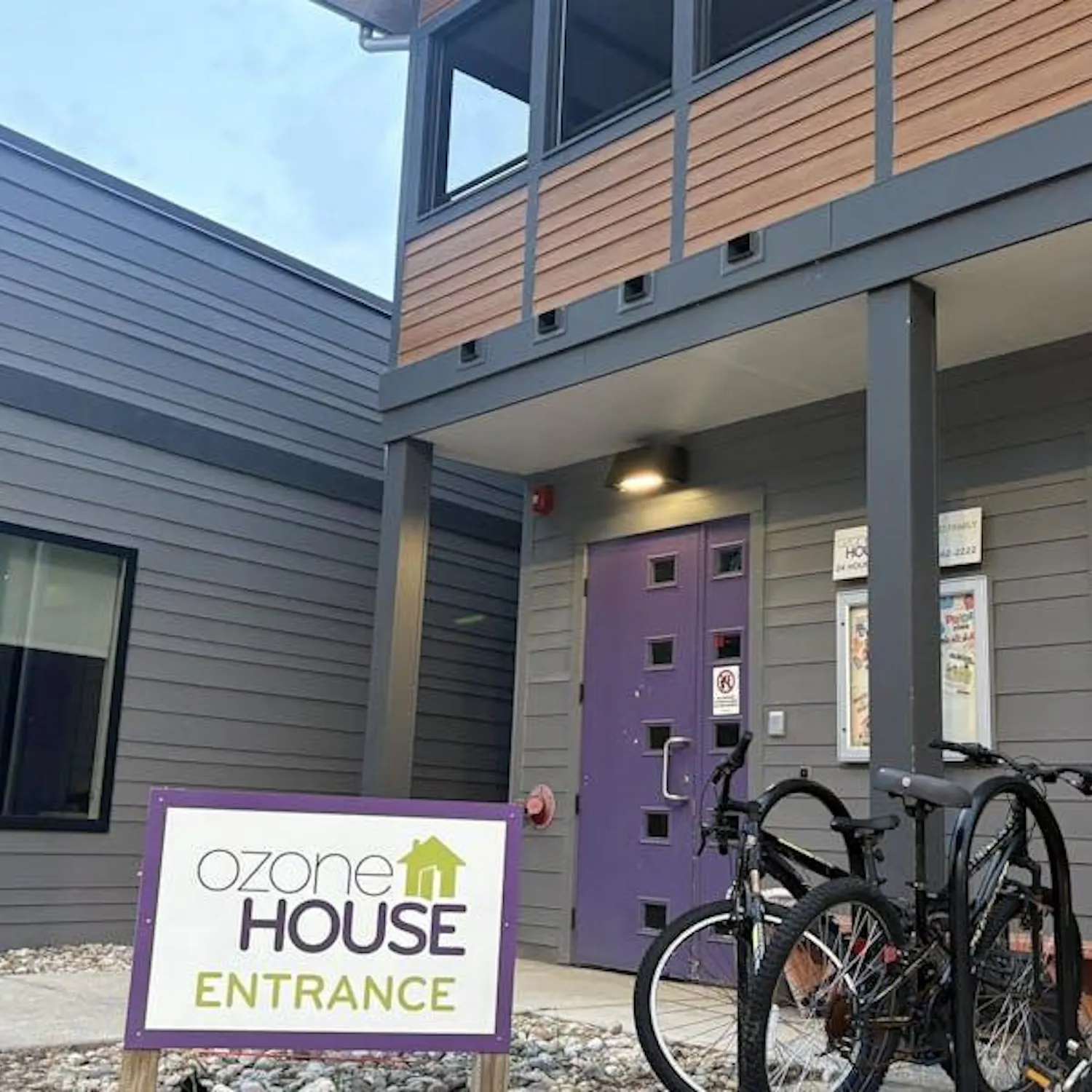 A square-shaped building made of gray-colored wood with a brown-colored panel running across its middle. There are several windows, and the door to the building is purple. In one of the windows is a Black Lives Matter sign. A few bikes are parked in front of the building, and a sign in the foreground says "Ozone House Entrance." Overhead, the sky is a shade of pale blue.