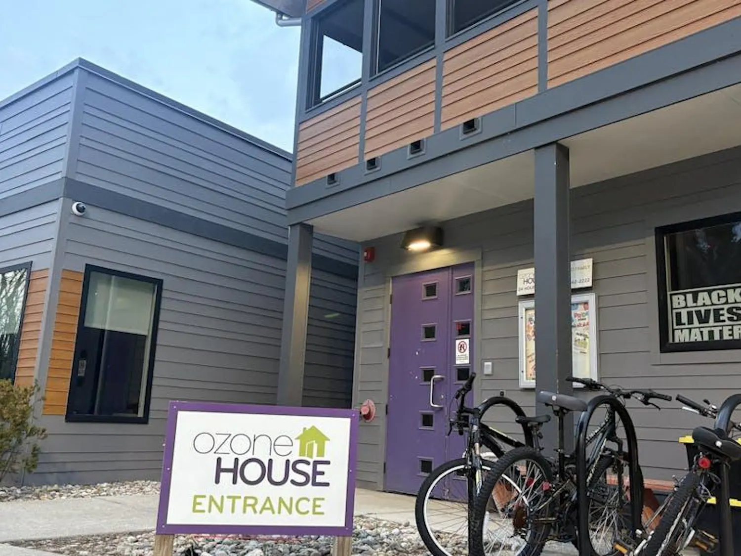 A square-shaped building made of gray-colored wood with a brown-colored panel running across its middle. There are several windows, and the door to the building is purple. In one of the windows is a Black Lives Matter sign. A few bikes are parked in front of the building, and a sign in the foreground says "Ozone House Entrance." Overhead, the sky is a shade of pale blue.