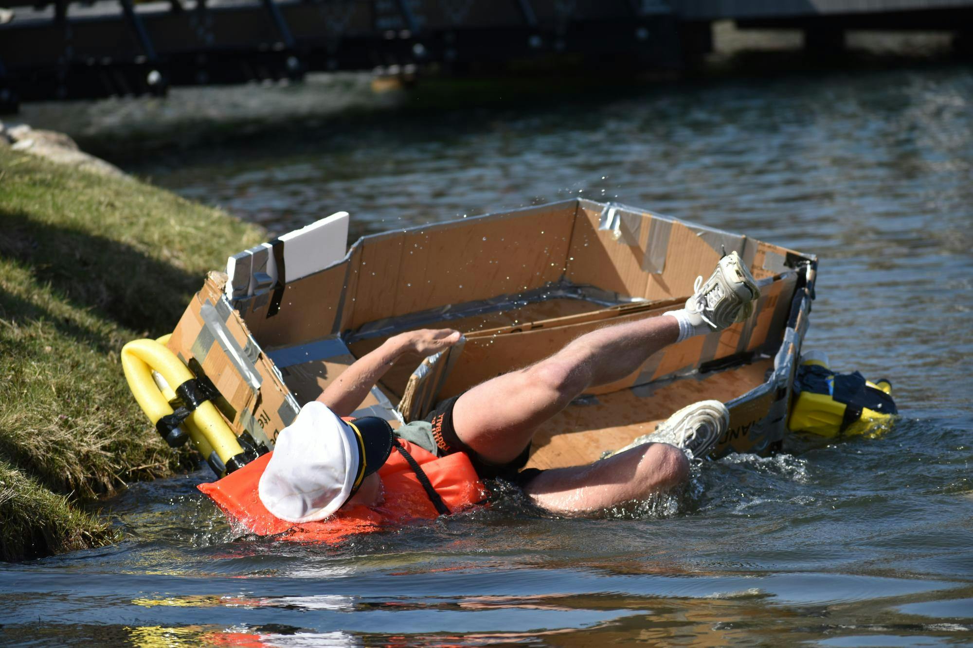 A student wearing a life jacket falling out of a handmade boat constructed with cardboard, plastic and tape, next to a grassy shore.