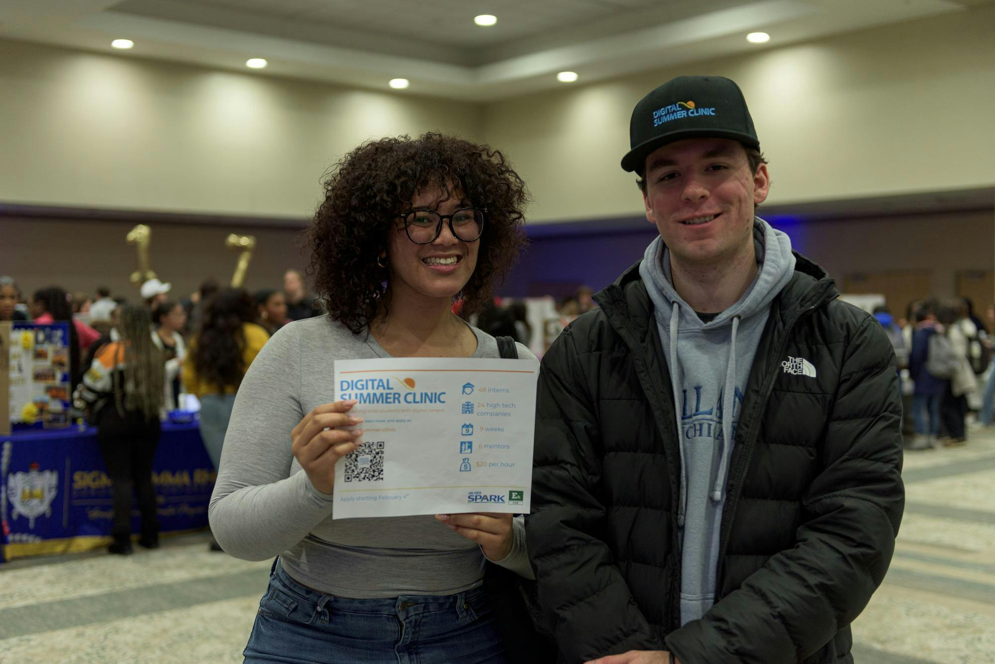 Two students stand next to each other and smile while holding a paper with information about Digital Summer Clinic. 