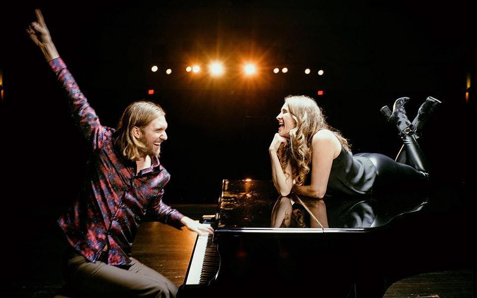 Drew gives a big smile while seated on the bench of a grand piano. Kin lies on top of the piano and smiles at Drew.