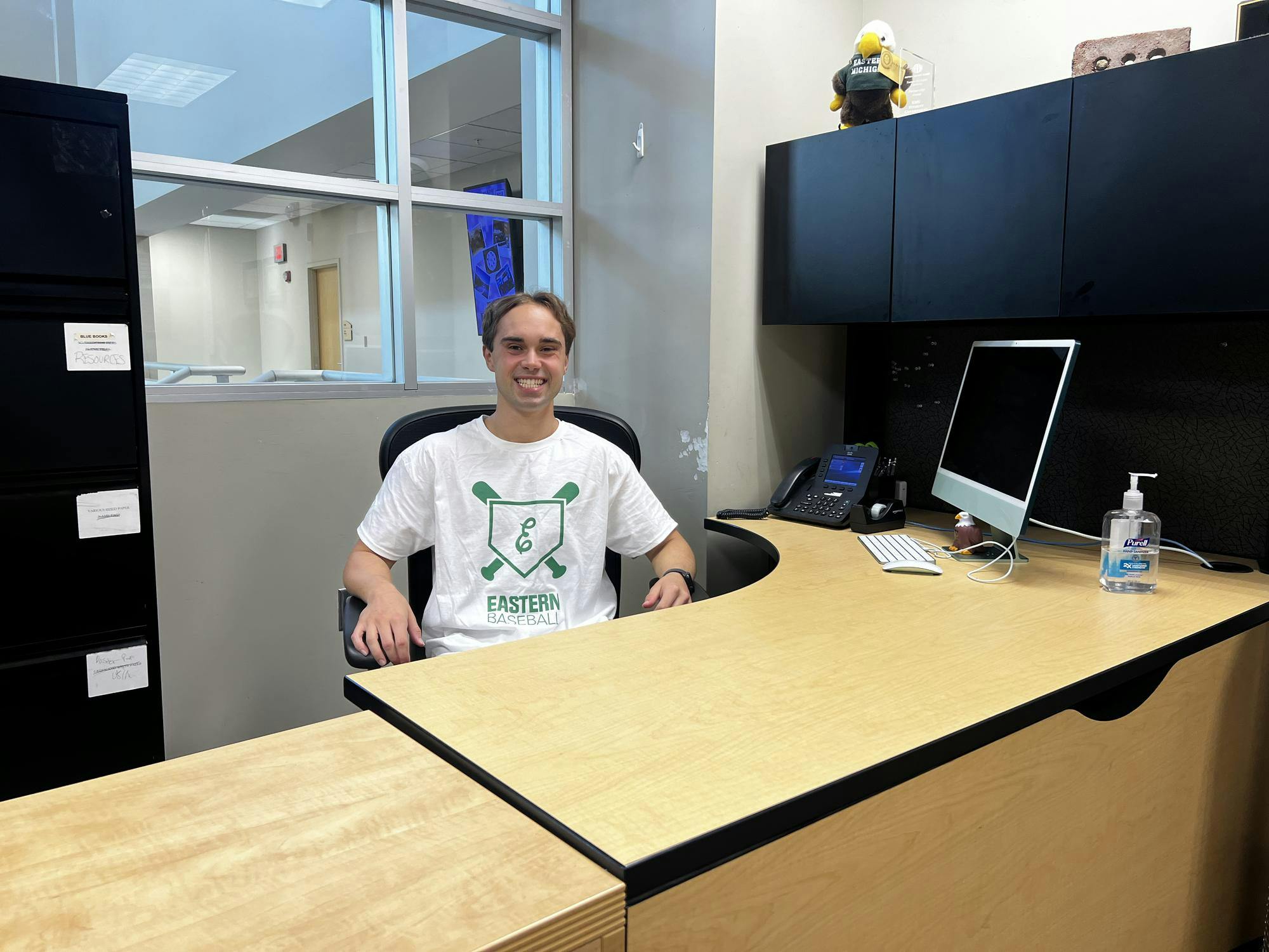 Jack Booth sits behind a wooden desk. He is smiling and wearing an Eastern Baseball shirt. A stuffed eagle "Swoop" plush sits on his cabinet. 