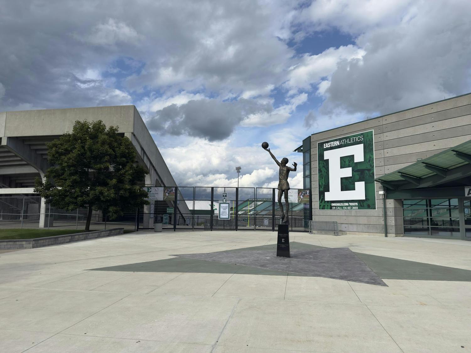 Gray cement sidewalks and structures surround the black gate, closed in front of a football stadium on a partly cloudy day. A metal statue of a basketball player holding a ball above its head is positioned in the middle of the walkway.
