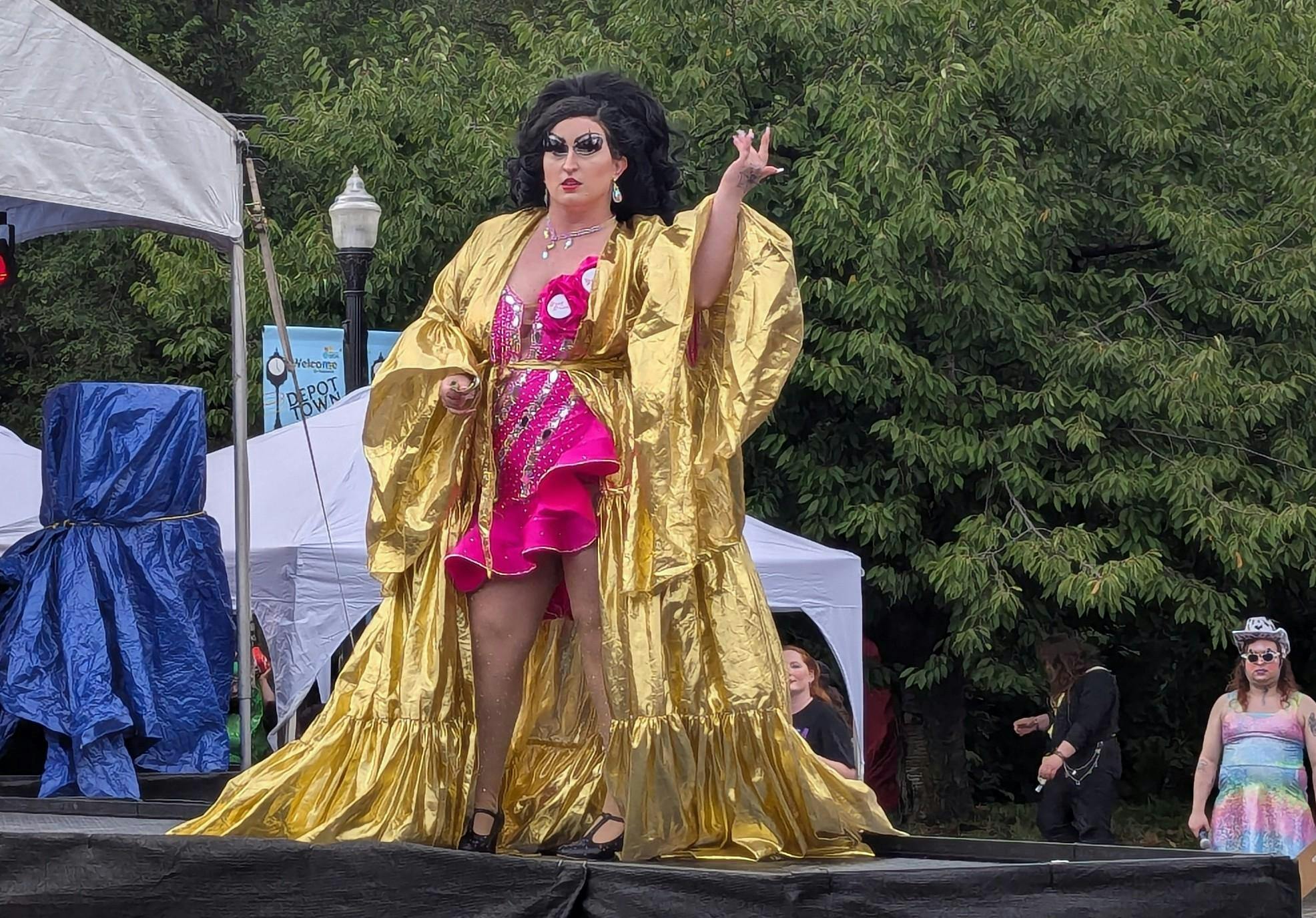 A drag queen in a flowing golden robe poses on stage mid-performance.