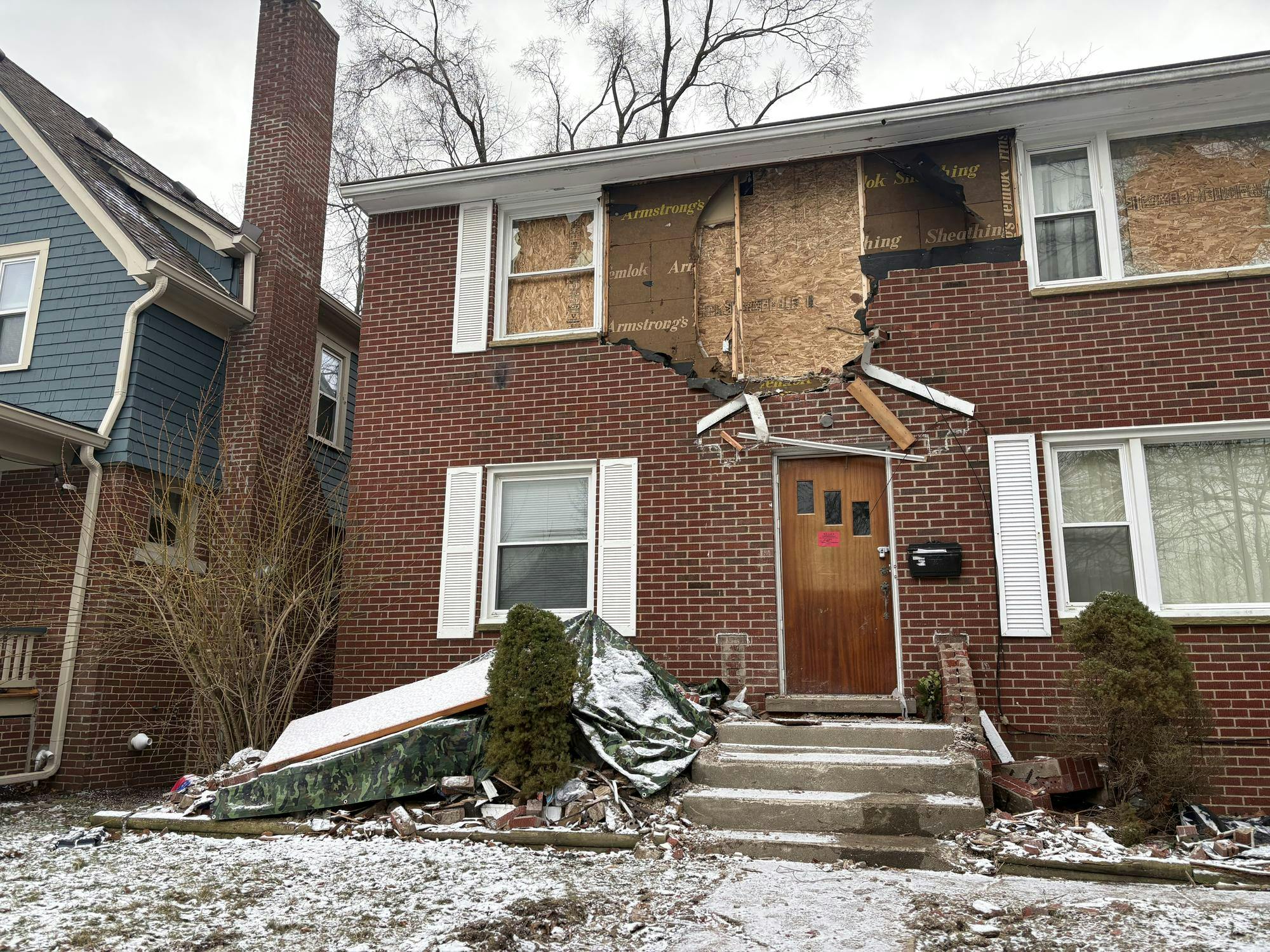 Two-story red brick house. 2 windows have been boarded up with plywood on the upper floor, along with a hole in the exterior brick wall between them. Tarps, bricks, and debris litter the front, snow-covered yard.