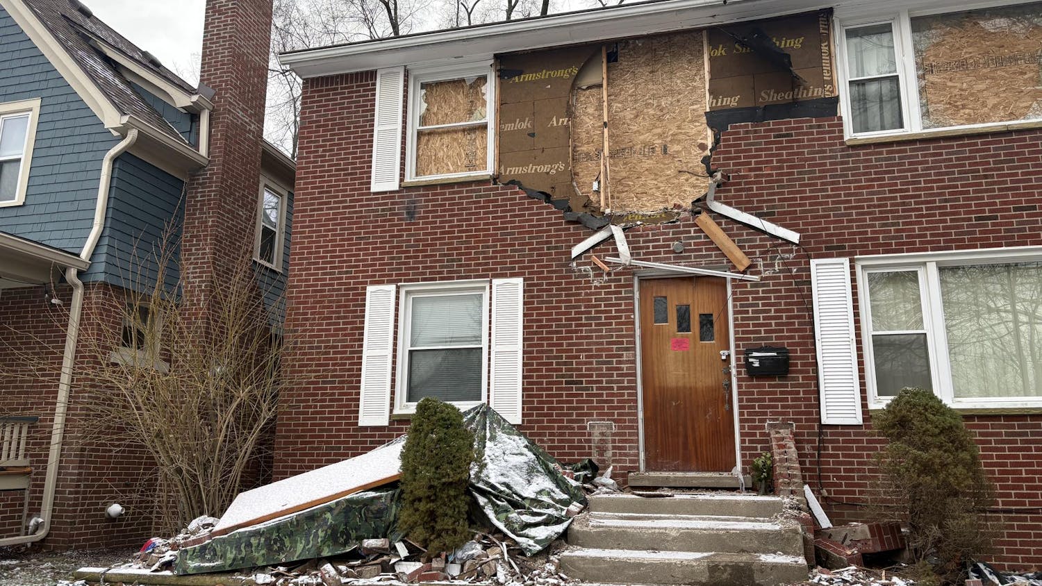 Two-story red brick house. 2 windows have been boarded up with plywood on the upper floor, along with a hole in the exterior brick wall between them. Tarps, bricks, and debris litter the front, snow-covered yard.