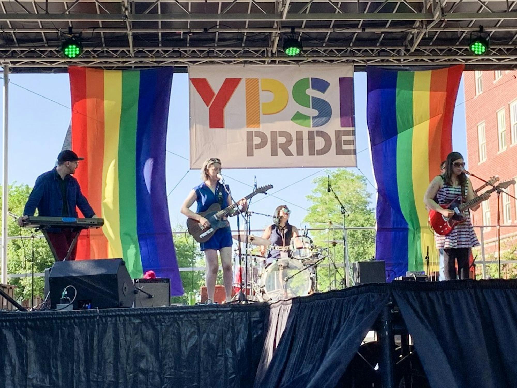A band of 4 individuals performs on a stage in front of an "Ypsi Pride" banner with rainbow lettering.