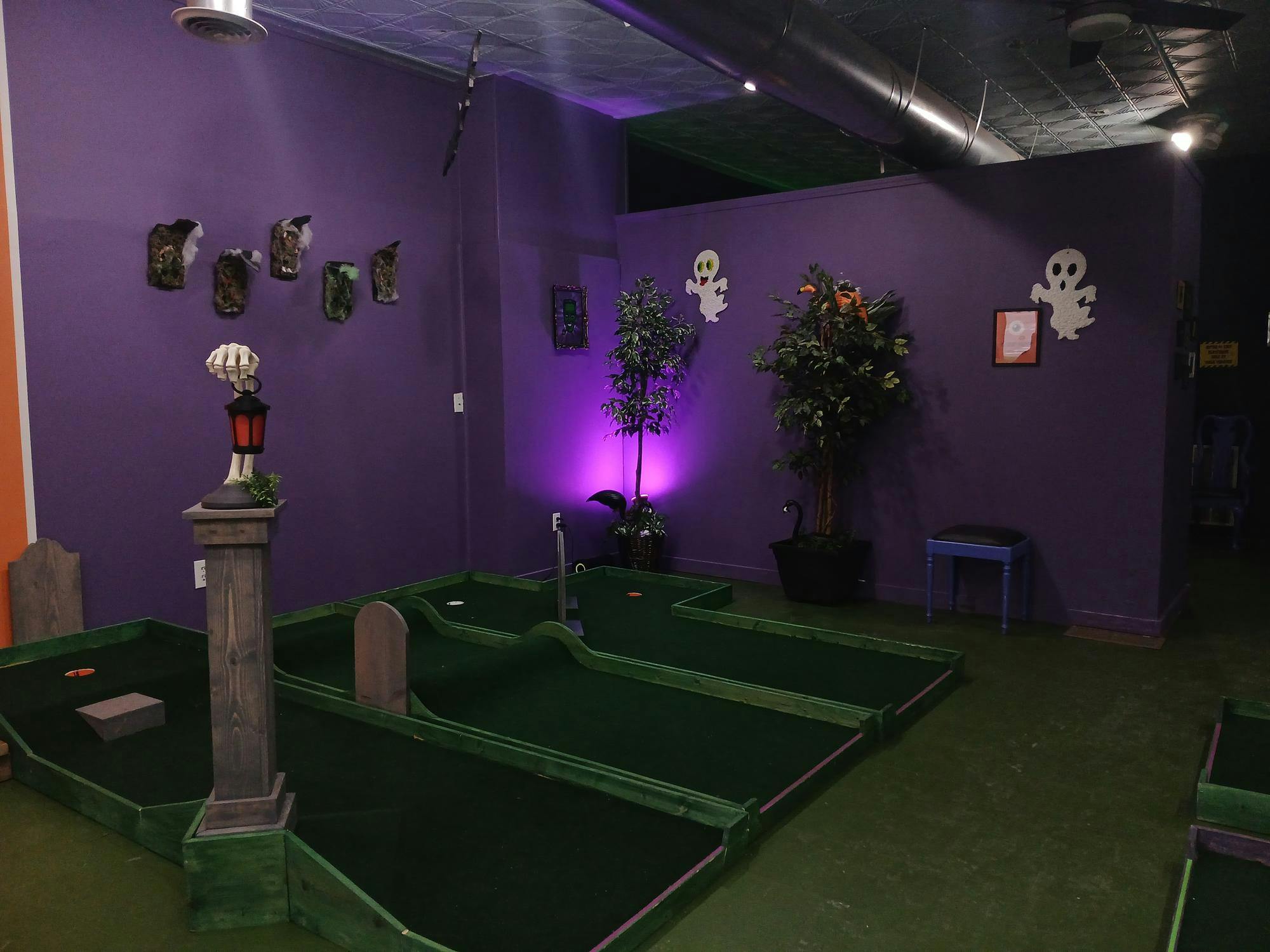 Purple walls in low lighting are decorated with ghost wall hangings and other spooky decor. There are three green indoor miniature golf course holes for playing on the ground.