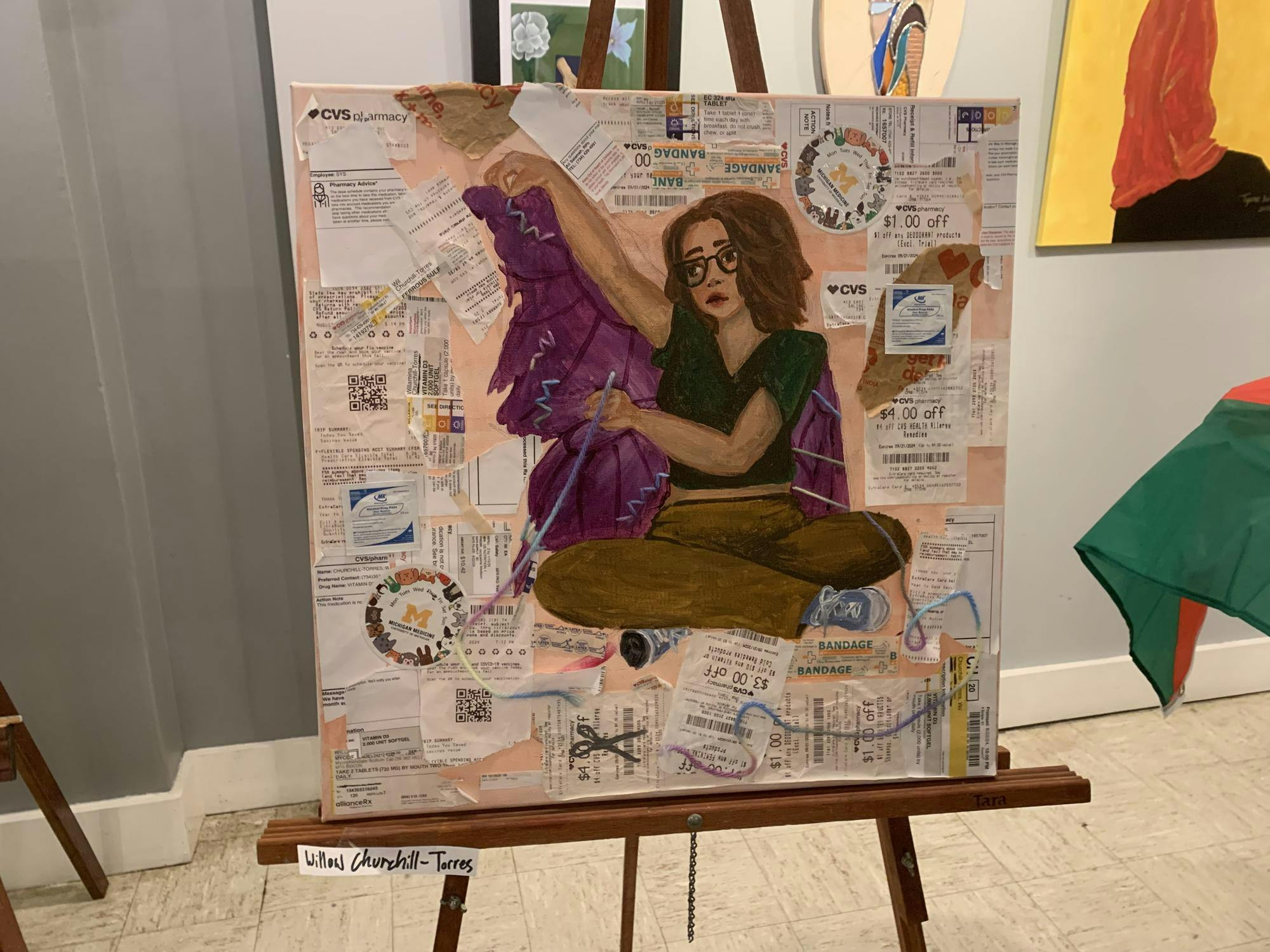 A person with glasses sits in the middle of a painting, surrounded by receipts from medical providers and pharmacy's. 