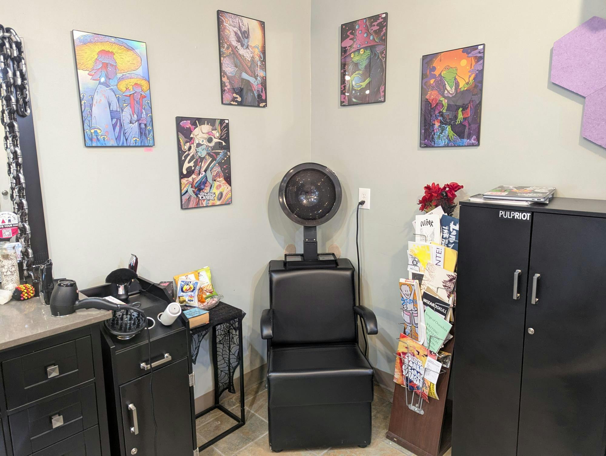 A salon hair-drying chair with local art displayed behind it and a rack of zines beside it.
