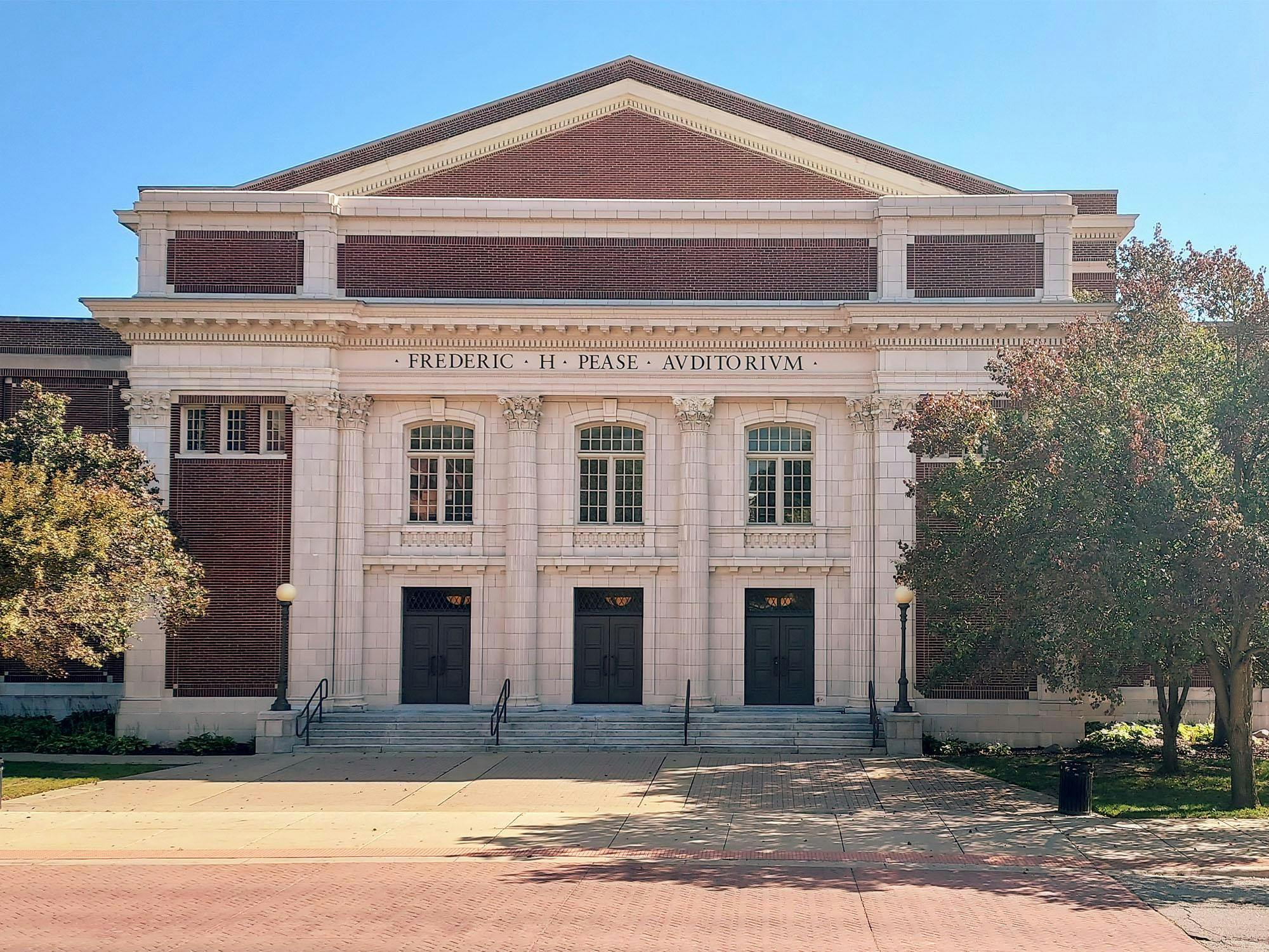 Pease Auditorium, a classical-style red brick and white cement building with three double doors and windows on the front. Corinthian columns decorate the front of the building.