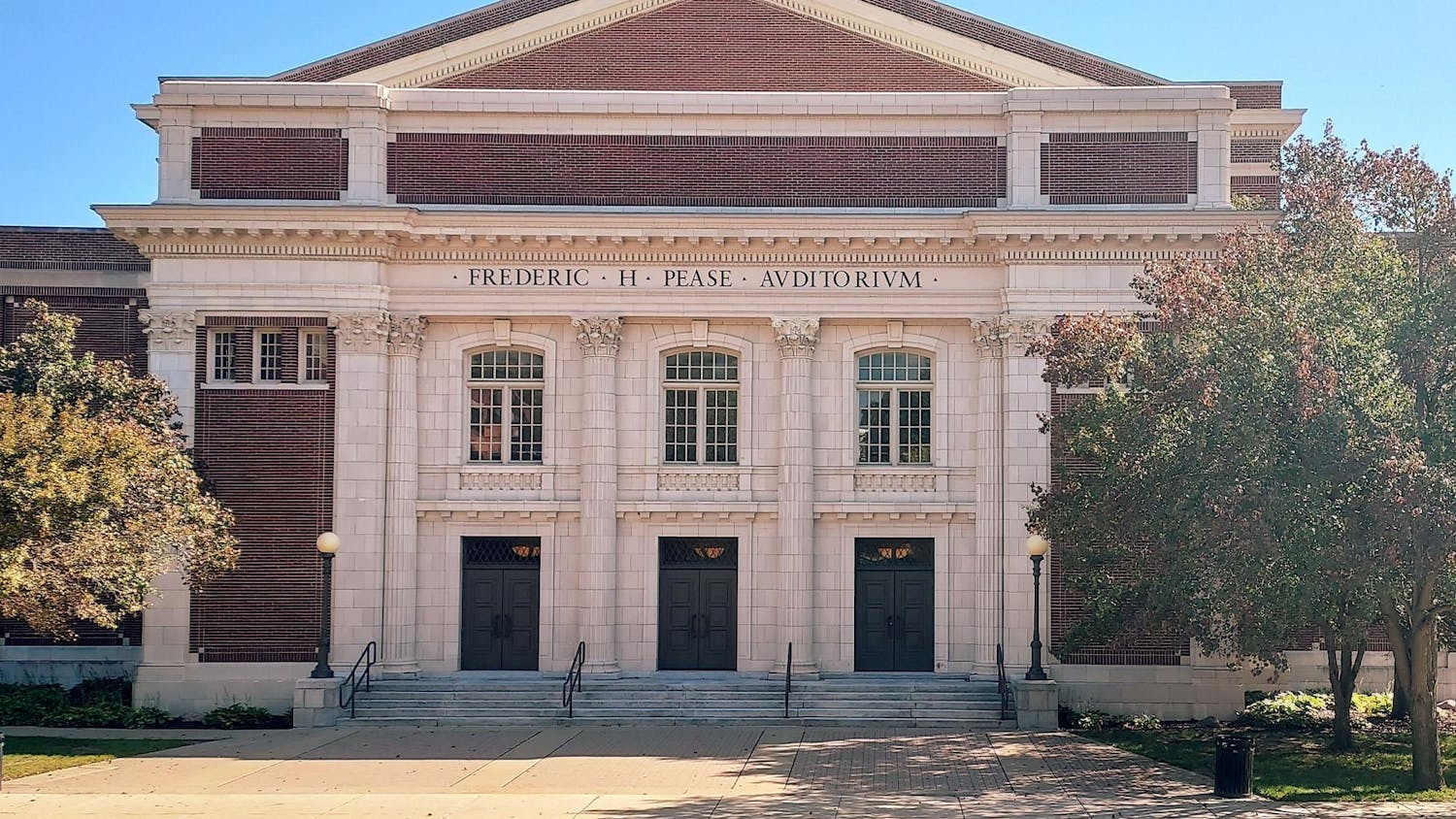 Pease Auditorium, a classical-style red brick and white cement building with three double doors and windows on the front. Corinthian columns decorate the front of the building.