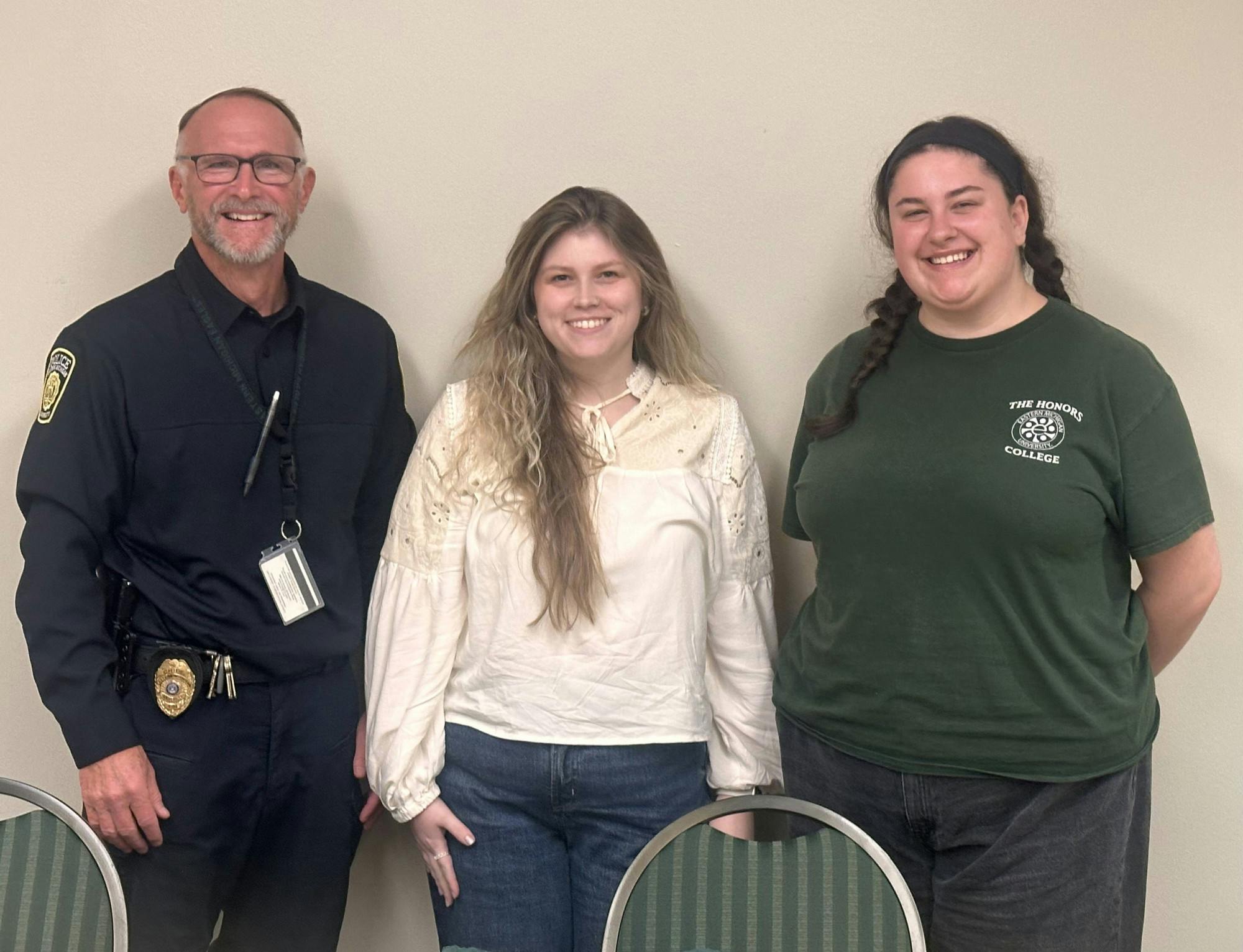 Chief Lige, in police uniform, Alyssa Mohr and Katie George stand smiling in front of a blank wall.