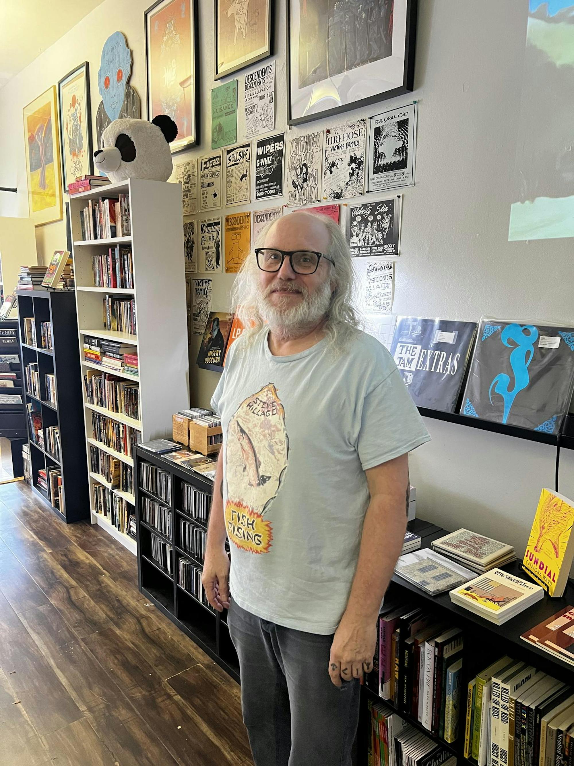 A man with long white hair and a beard standing in a record store wearing a light blue Steve Hillage shirt and glasses.