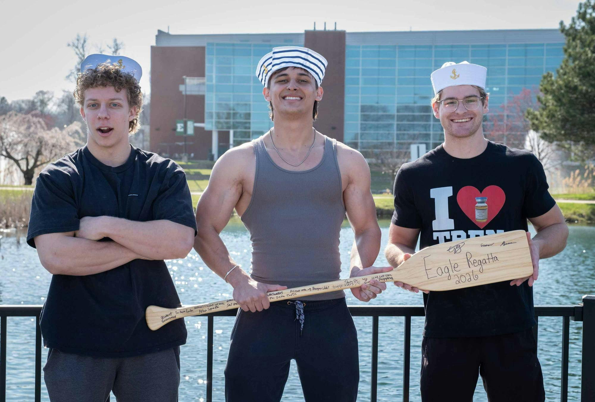 Three students stand in front of a pond smiling and holding a wooden oar with writing that serves as the trophy for the 2026 Eagle Regatta.