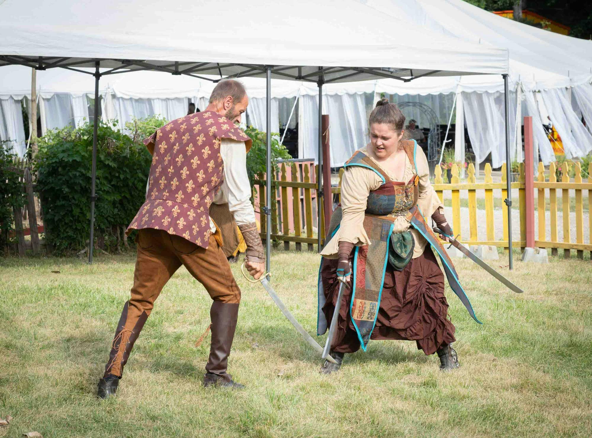 Two people dressed in medieval clothing sword fight outdoors.