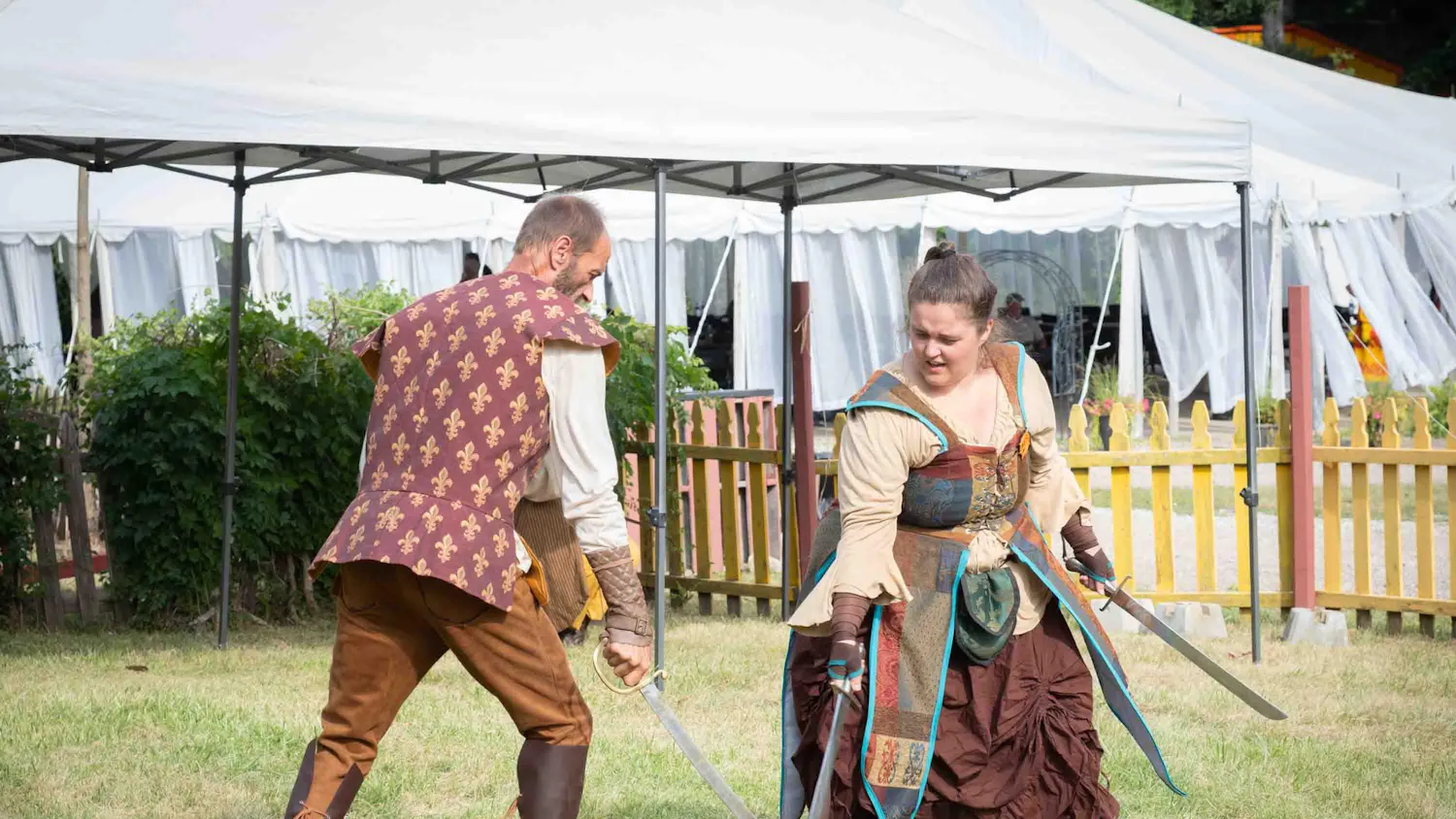 Two people dressed in medieval clothing sword fight outdoors.
