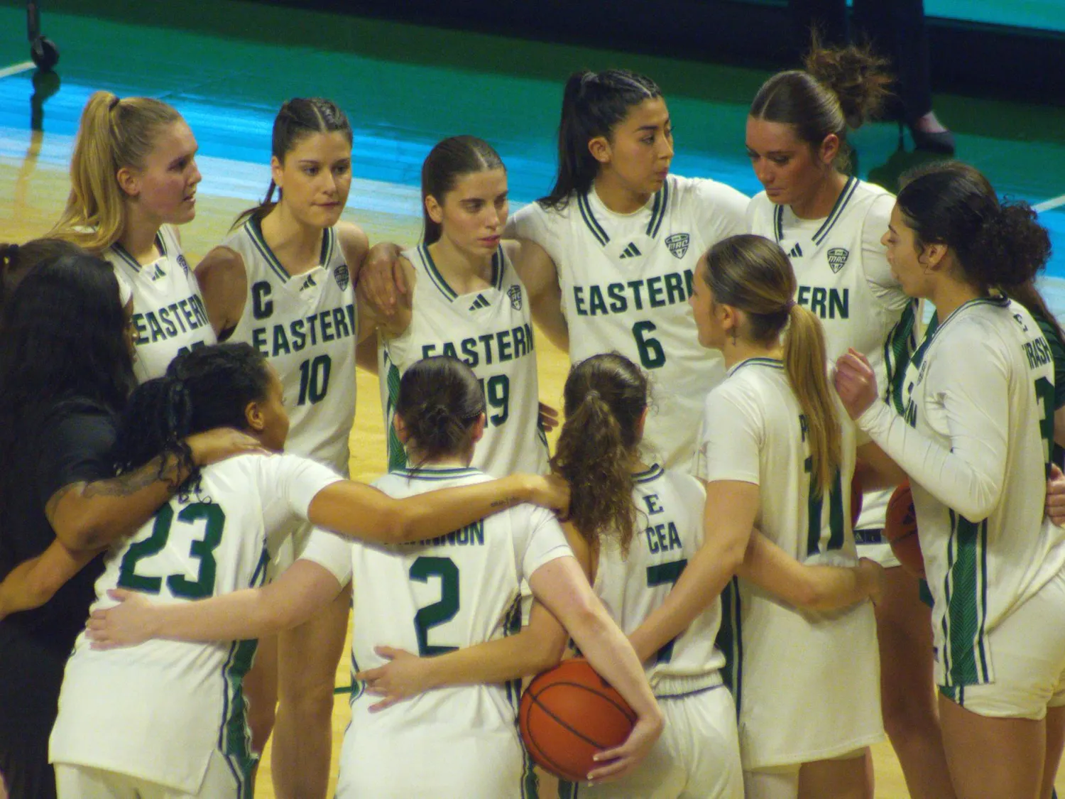 Women's basketball players stand in a ring wearing white EMU jerseys with green numbers and text.