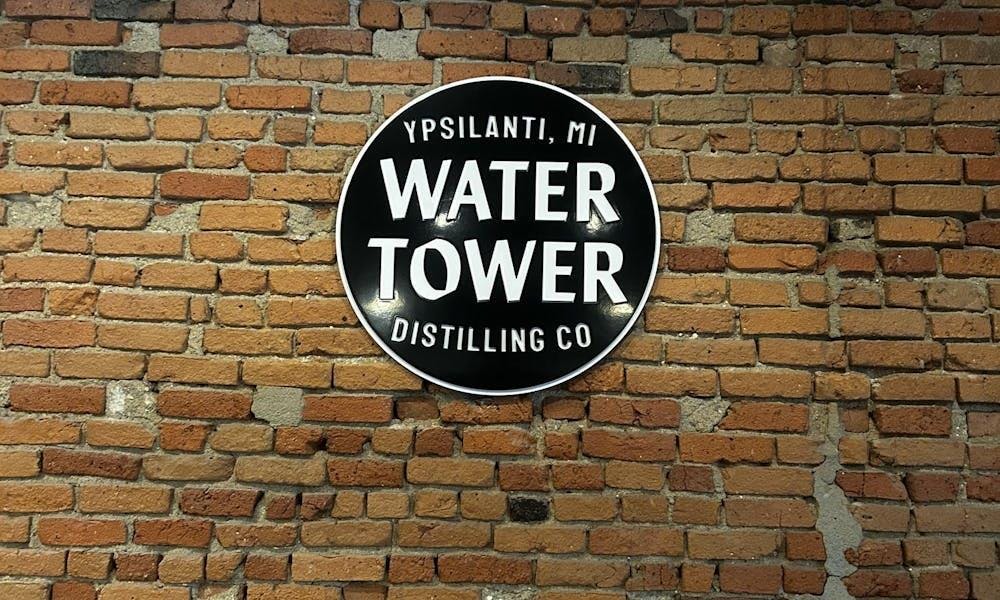 A red brick wall with a circular black sign hanging on it. There are four lines of white print on the sign. The first says "Ypsilanti, MI." The second says "Water," the third says "Tower," and the fourth says "Distilling Co." All of the letters on the sign are capitalized.