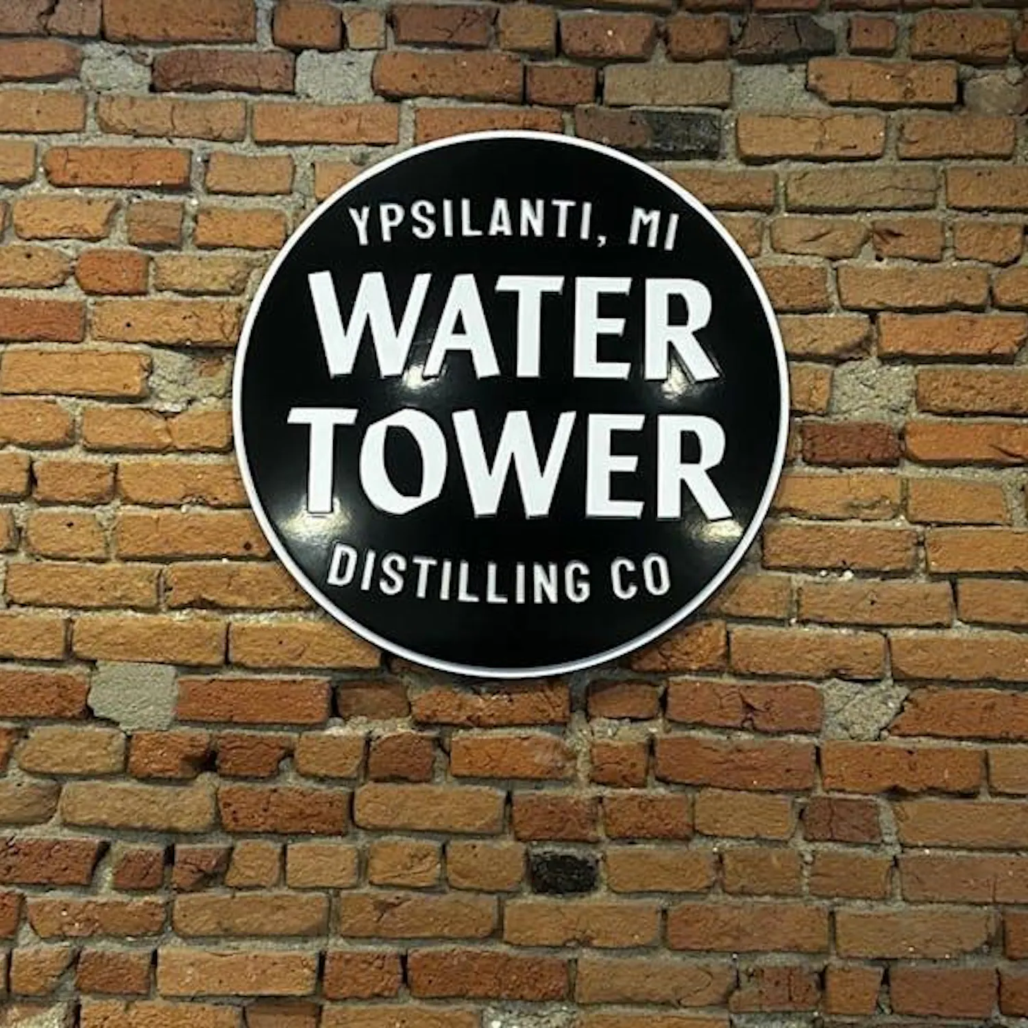 A red brick wall with a circular black sign hanging on it. There are four lines of white print on the sign. The first says "Ypsilanti, MI." The second says "Water," the third says "Tower," and the fourth says "Distilling Co." All of the letters on the sign are capitalized.